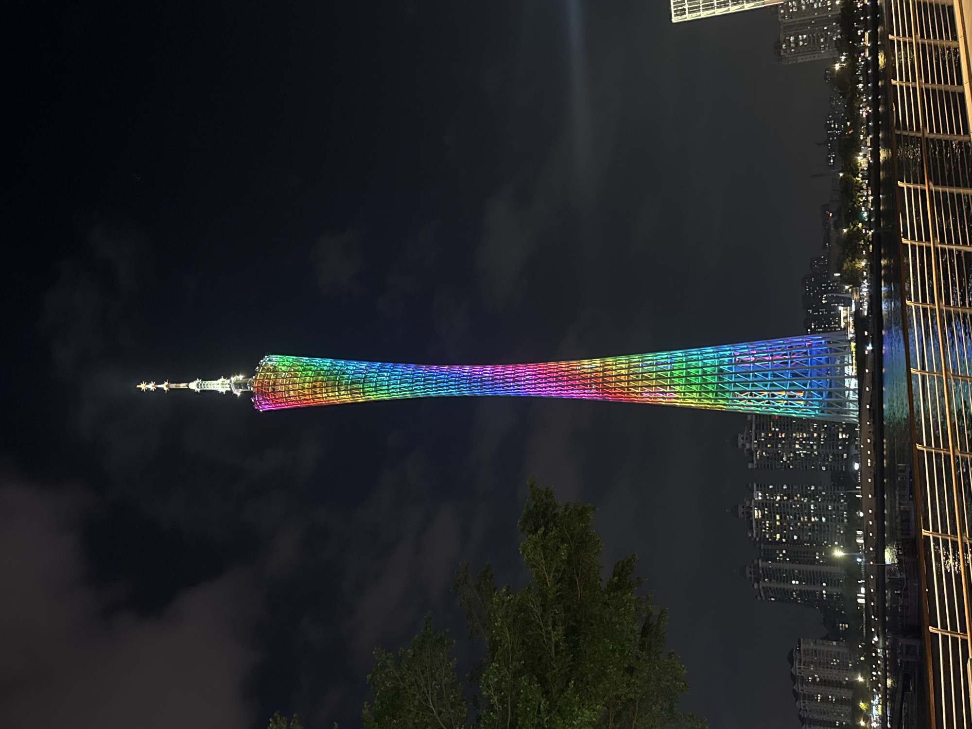 The image shows the Canton Tower at night, located in the Tianhe District, China. The tower is illuminated with a rainbow of colors. There are apartment buildings to the right side of the image. There are trees in the foreground.