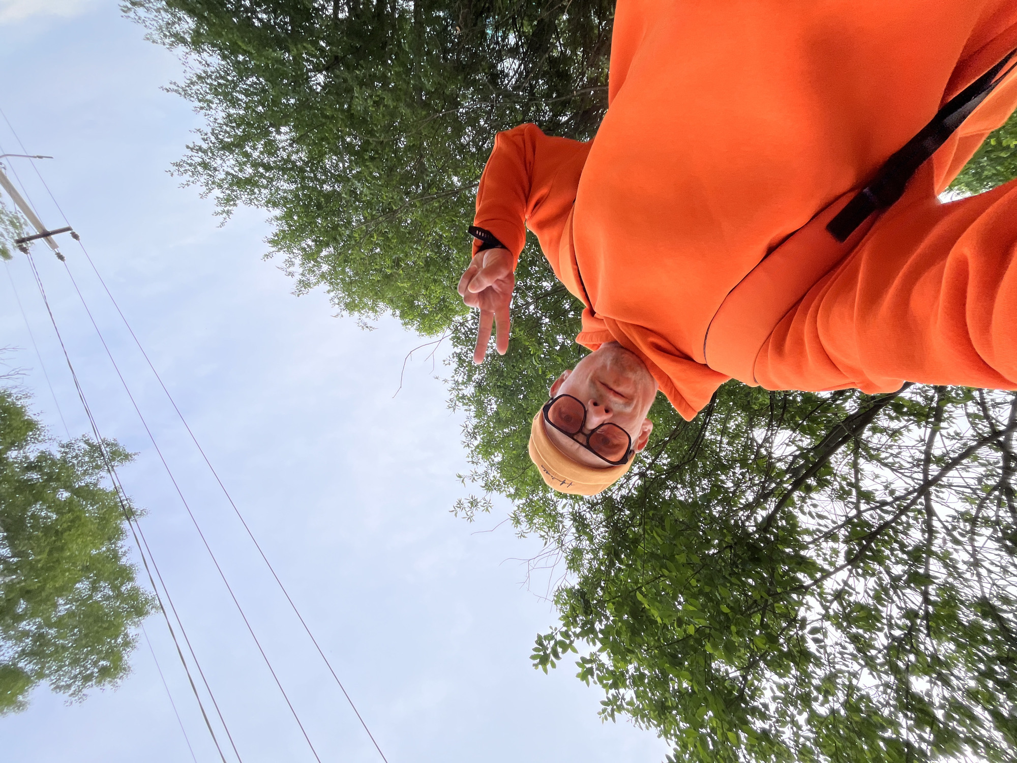 A low-angle, upward-looking photograph captures a person striking a pose against a backdrop of the sky and tree canopy in Chaikovsky, Russia. The individual, positioned upside down relative to the ground, occupies the upper half of the frame. They are wearing a vibrant orange hoodie or sweatshirt, a light-colored beanie or cap, and amber-tinted sunglasses. The person's left hand is raised, forming a "peace" sign with two fingers, directed towards the camera.
The background features a light blue sky, suggesting daylight conditions, with sparse, wispy clouds. Lush green leaves of deciduous trees fill the right and top portions of the image, indicating a natural outdoor setting. On the left side, a utility pole with multiple power lines is visible, partially obscured by some tree branches. The overall impression is an informal, outdoor portrait taken with a unique, playful perspective. No visible text is present in the image.