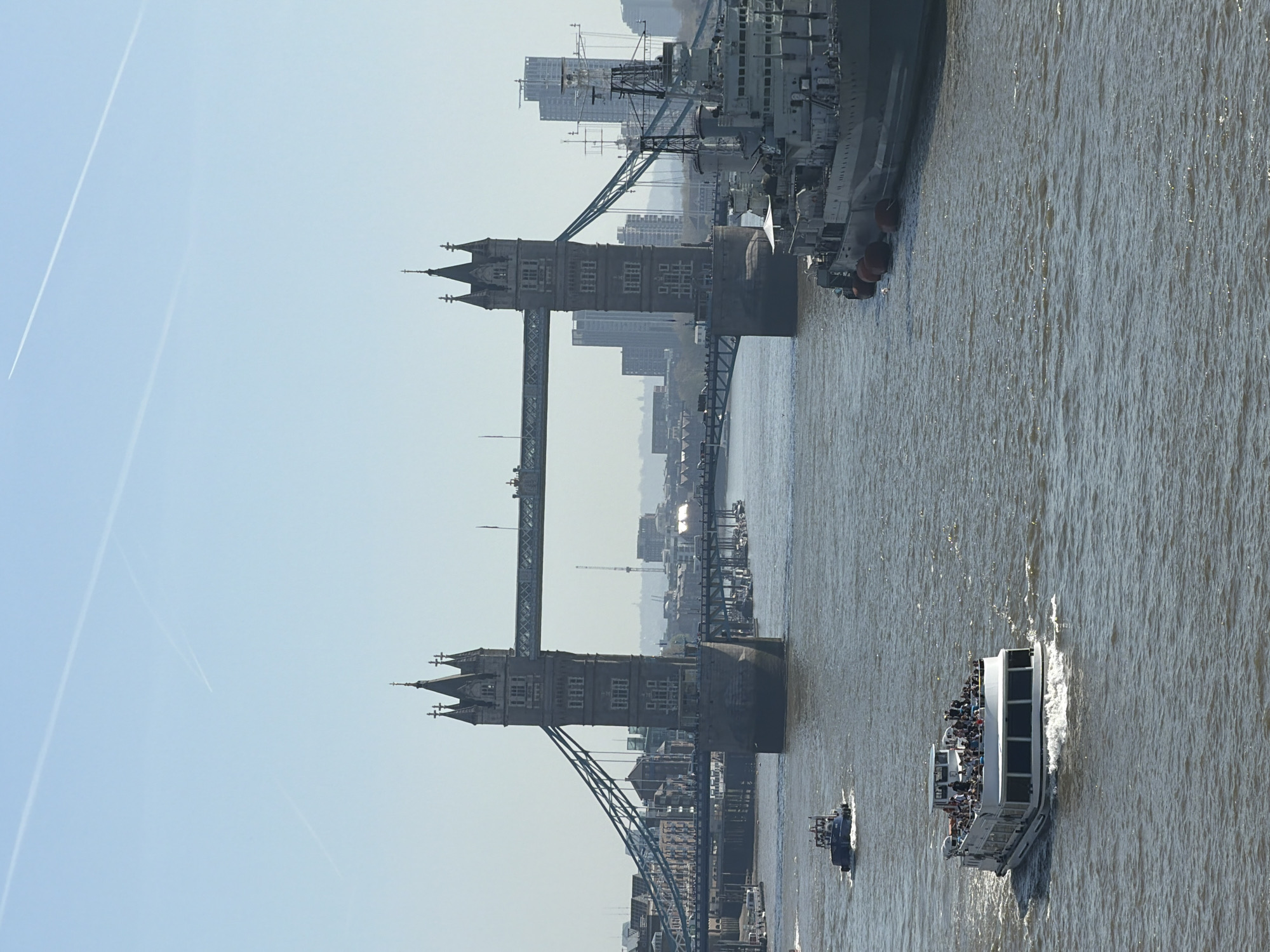 The image depicts the Tower Bridge in London, United Kingdom, spanning the River Thames. The bridge's iconic towers are visible, connected by the elevated walkways and the bascules. Several boats, including a large passenger ferry, are on the river. A large ship is docked alongside the bridge, filling the gap between the bridge towers. The sky is clear, with contrails of an aircraft visible. The overall environment suggests a bright, daytime setting. The water appears calm. The details of the surrounding buildings are partially obscured by the haze and distance.