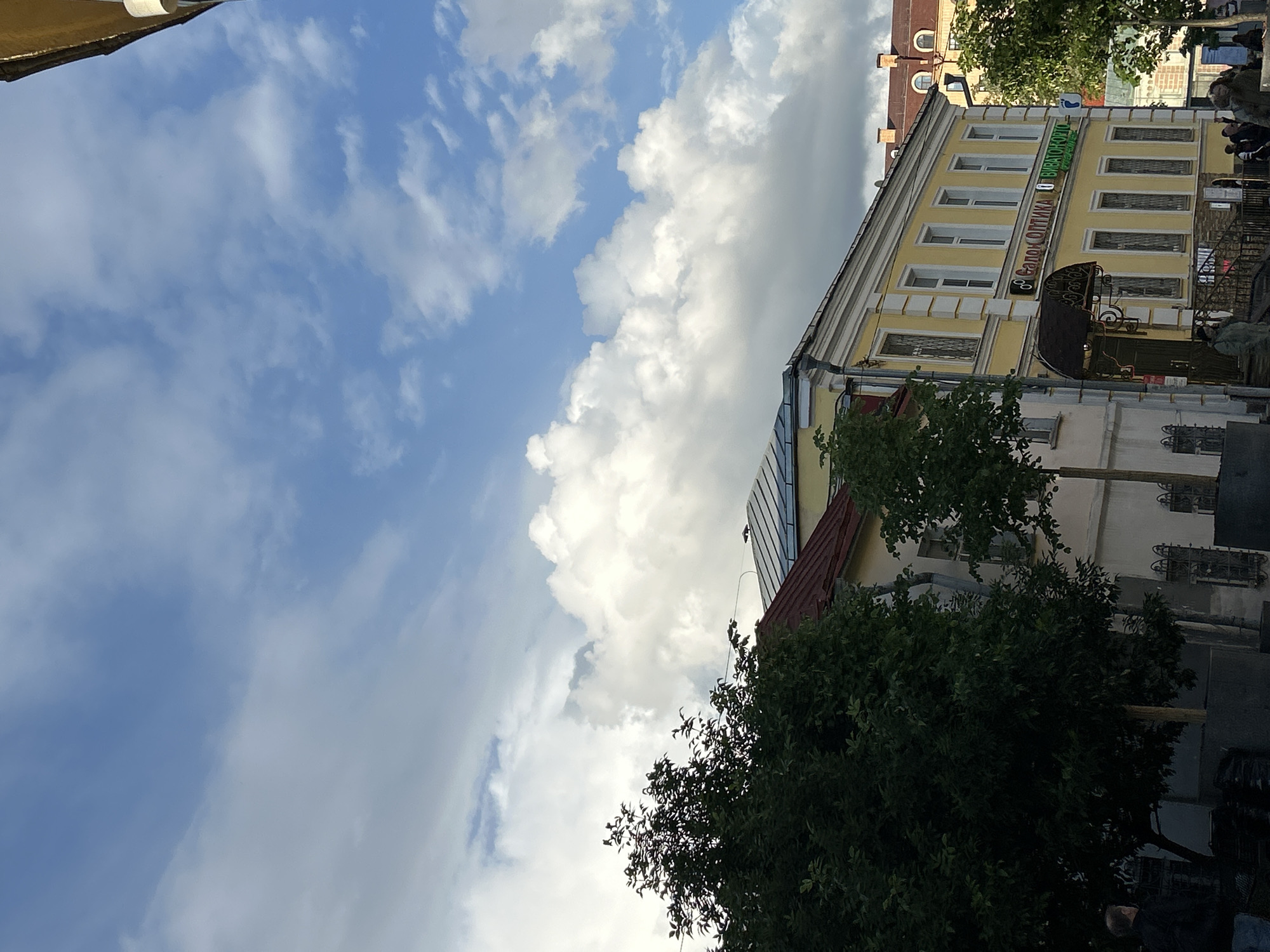The image shows a view of a building in an urban setting, likely a city. The sky occupies a significant portion of the frame, filled with a mix of light blue sky and large, puffy white and gray clouds.
The building on the right side of the image is multi-storied, featuring a facade with windows and architectural details. A sign with Cyrillic script "Балок оптика" is visible. There are trees with dense green foliage near the building.
Several people are visible on the upper right side of the image. Their faces are not recognizable.