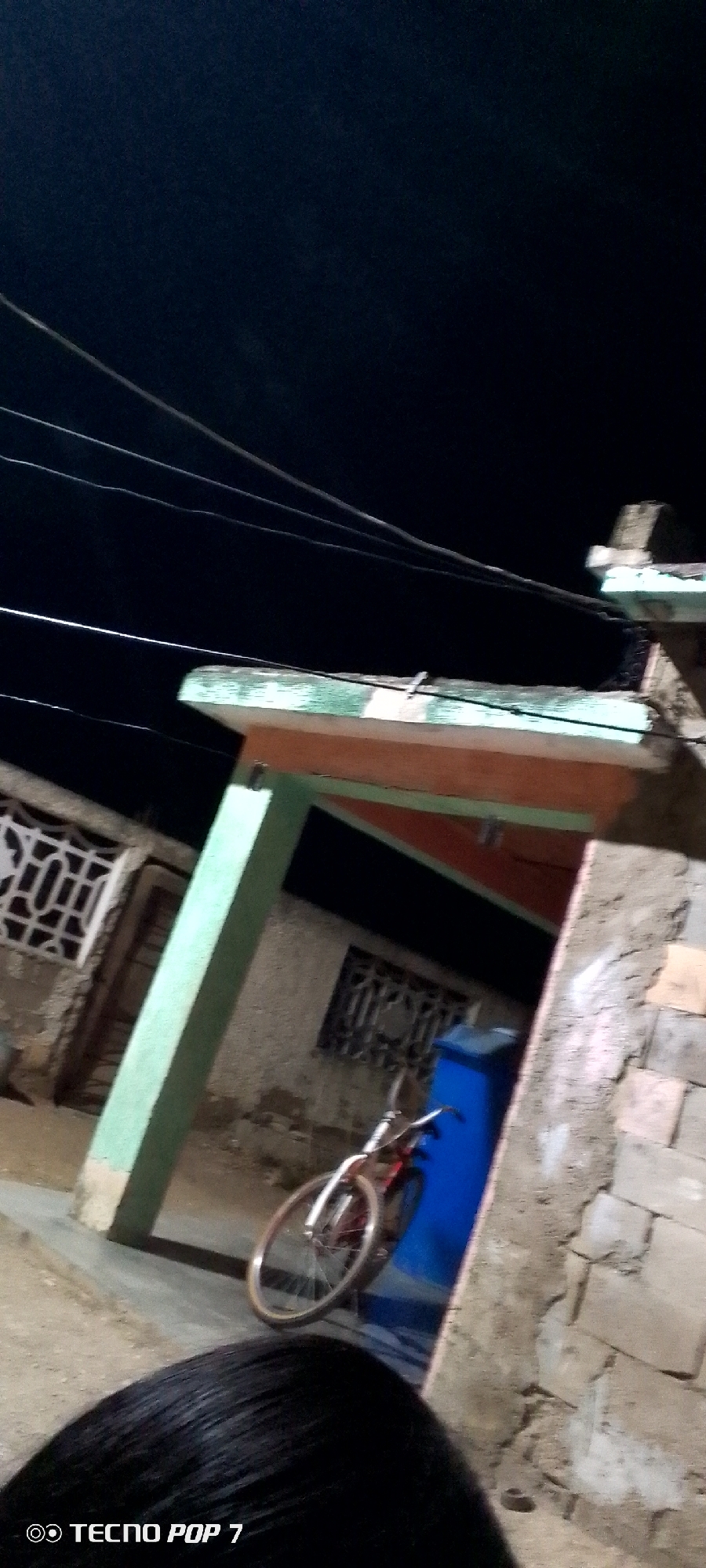 The image, captured in what appears to be Altagracia, Venezuela, shows a nighttime scene outside a building. The background is dark, with the sky mostly obscured. Overhead power lines are visible against the night sky. The building has a simple architectural design with a light green pillar. The exterior walls show signs of wear and age. In front of the building, a bicycle stands beside a blue object, possibly a container. The bottom of the image shows a person's hair. The overall lighting is poor.