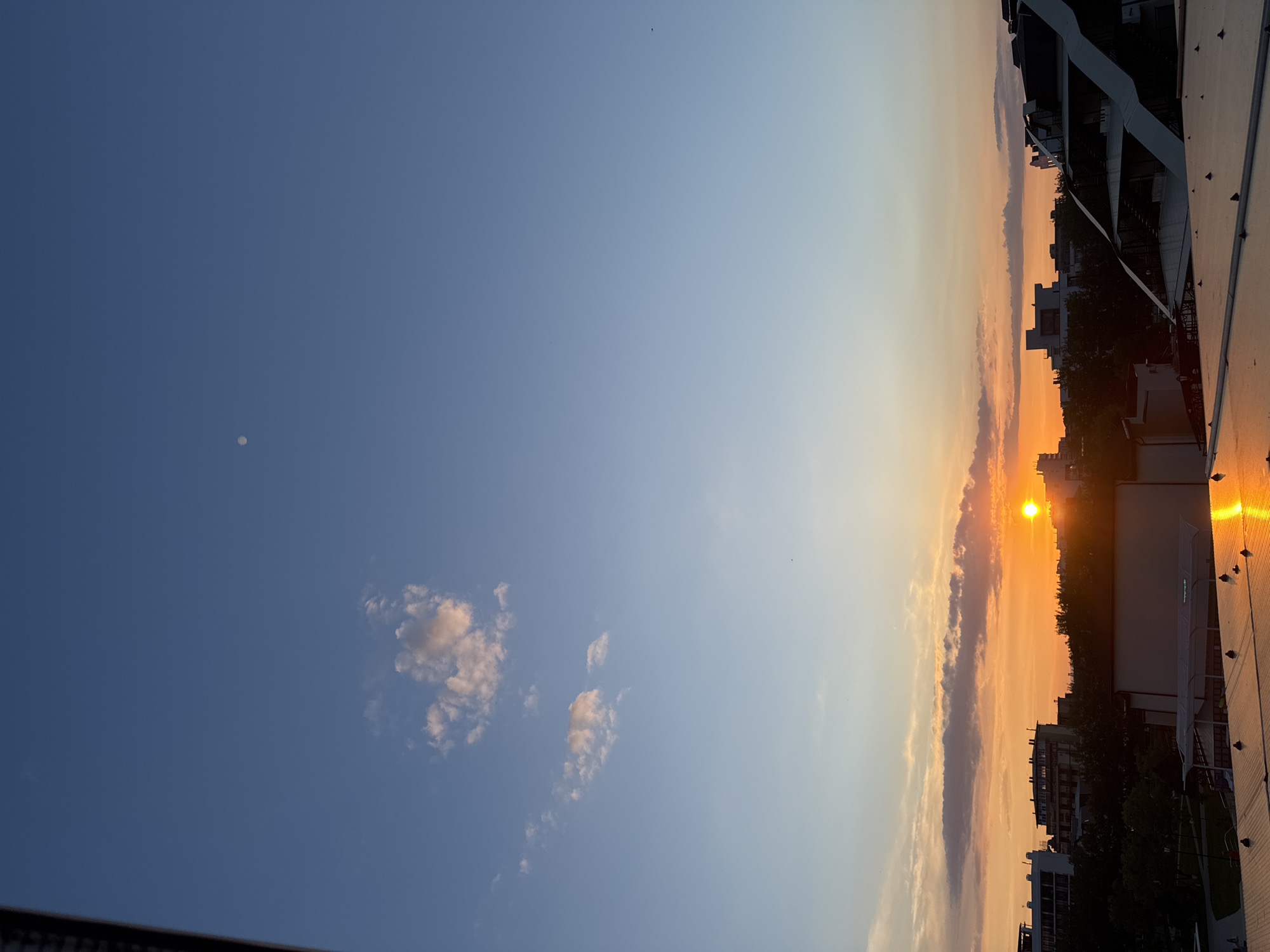The image depicts a sunset over a city, likely Vityazevo, Russia. The sky is a gradient of blue, transitioning to orange and yellow hues near the horizon where the sun is visible. Several clouds are scattered across the sky, some tinged with the colors of the sunset. In the distance, the silhouettes of buildings and trees form the cityscape. A bright reflection of the sun is cast on the rooftop in the foreground, which appears to be made of metallic material and has visible fasteners. There are no people or animals clearly discernible in the image. The overall scene captures a serene evening ambiance.