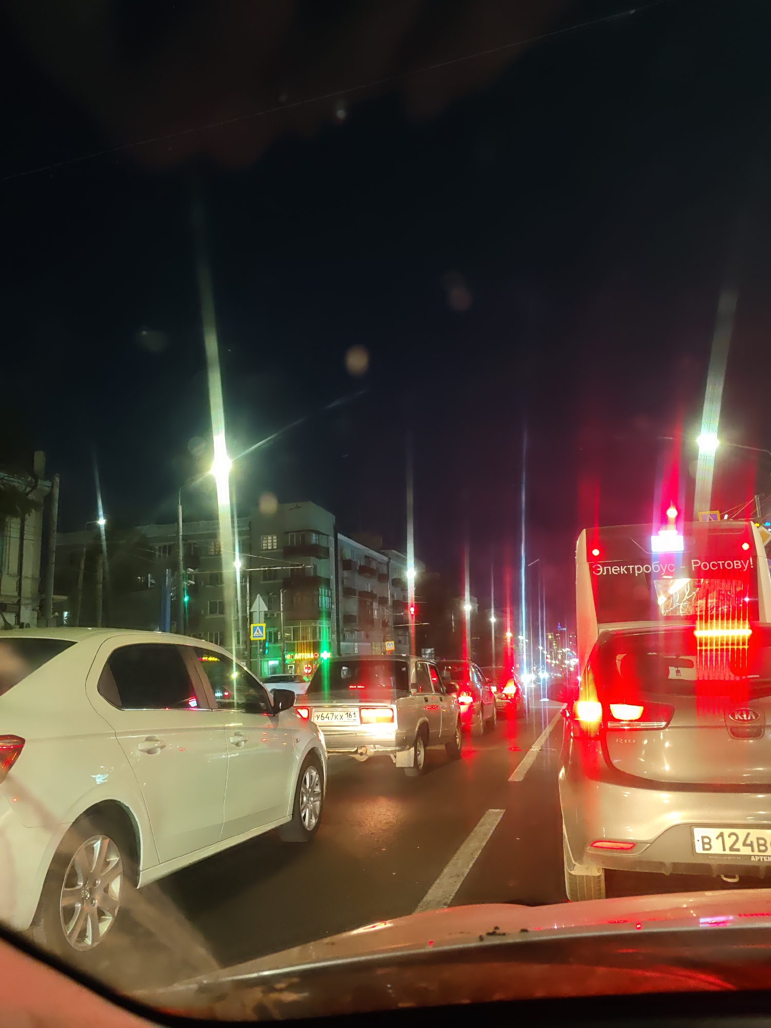 The image depicts a night-time traffic jam in Rostov-on-Don, Russia. The perspective is from inside a vehicle, looking forward. Several cars and a bus are visible, all stopped in traffic. The bus in the foreground has "Электробус - Ростову!" (Electric Bus - To Rostov!) written on its rear. The street is illuminated by streetlights and vehicle lights, creating reflections on the wet road surface. Buildings line the street on the left side. The overall scene is one of urban congestion at night.