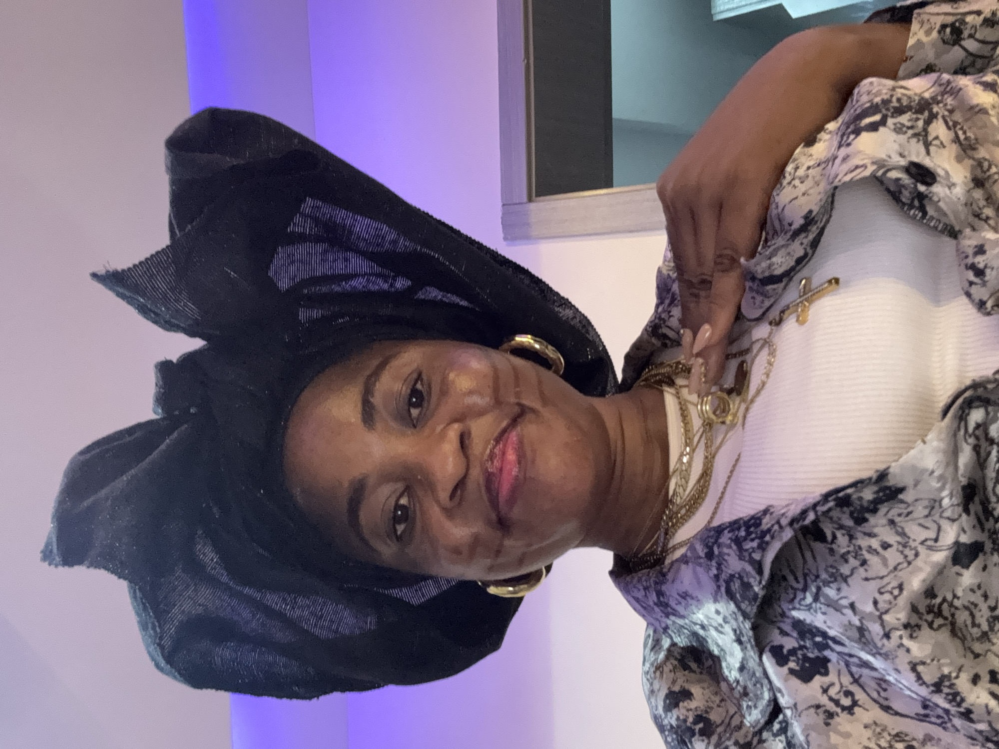 A woman wearing a black headwrap and gold hoop earrings poses for a selfie. She is also wearing a white ribbed top and a patterned jacket. Multiple gold necklaces are layered around her neck, one with a cross pendant. Her right hand is resting on her chest, with manicured nails. The background is a softly lit room with a purple hue, suggesting it might be a living space or a social gathering. The image was taken in Ajara, Nigeria.