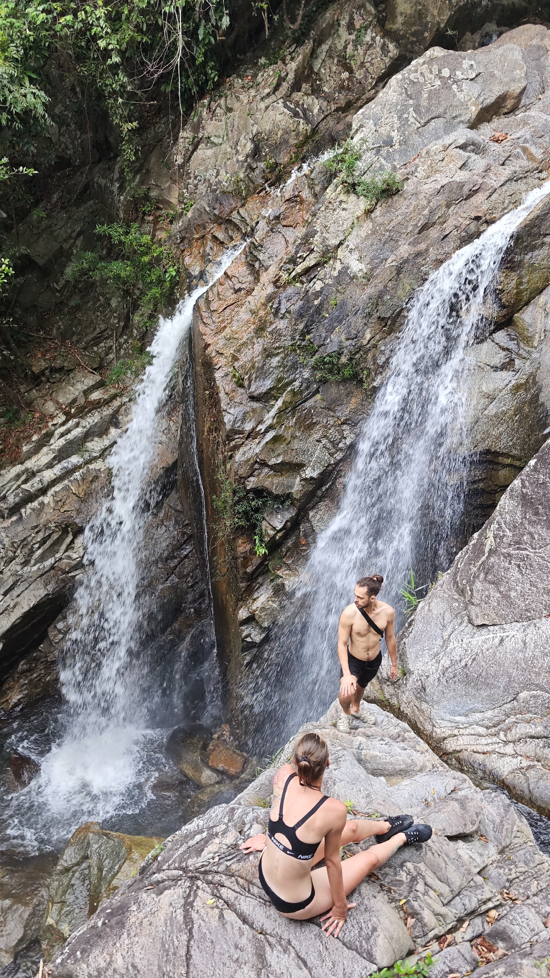 The image depicts a rocky waterfall environment, possibly in a natural area near Đà Nẵng, Vietnam. Two individuals are present: a woman sitting on a large rock in a black bikini and black athletic shoes, and a man standing on another rock in black shorts and a black shoulder bag. Two waterfalls cascade down the rocky terrain, with lush green vegetation visible at the top. The woman is looking away from the camera while the man looks directly ahead, seemingly into the waterfall. The rock formations appear rough and textured. The overall lighting is bright, with the sun illuminating the scene.
