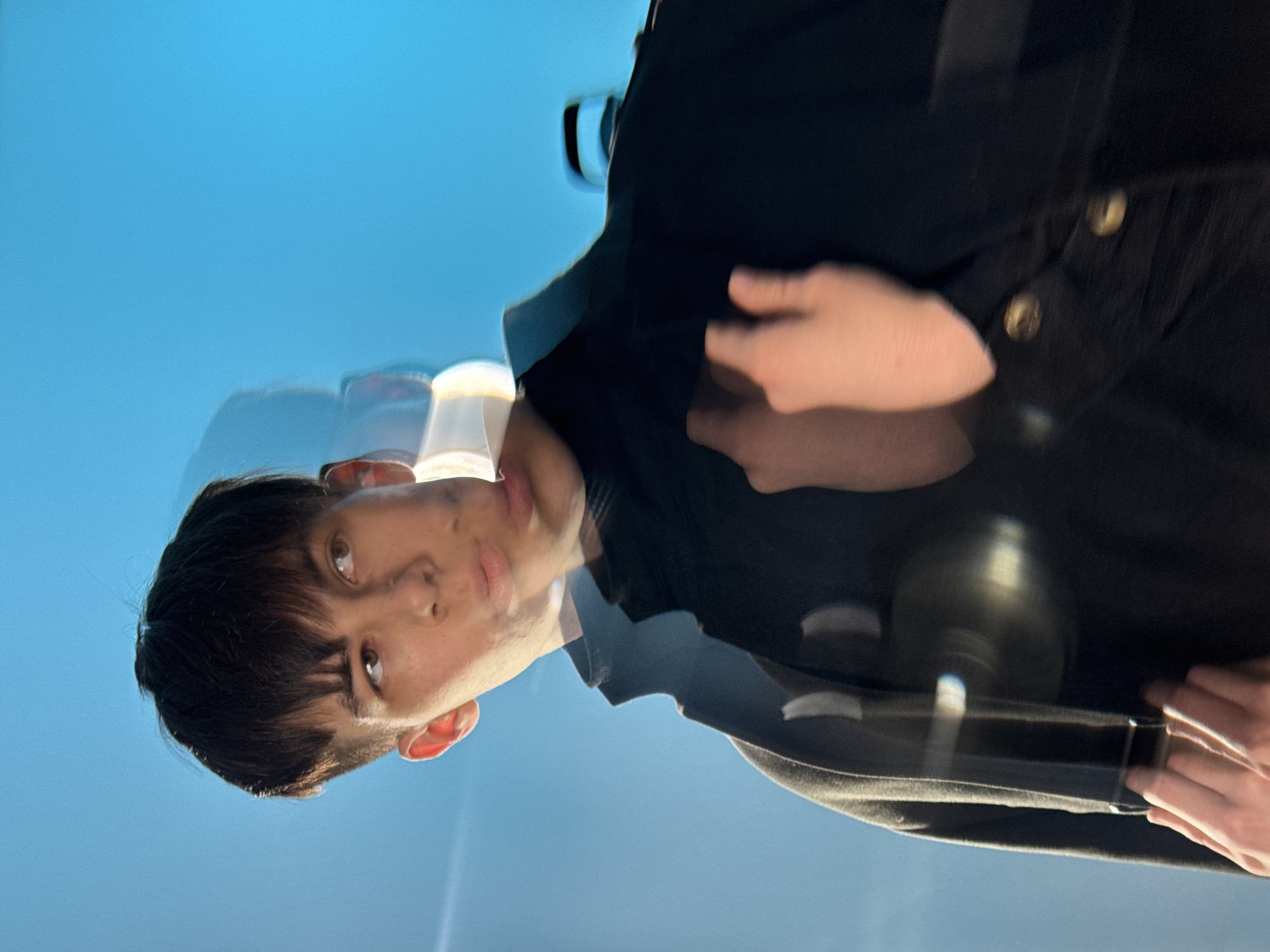 A blurred image depicts a young man with short, dark hair, facing slightly upwards. He has fair skin and dark eyes. He's wearing a dark jacket or shirt with buttons visible on the right side of the frame. The background is a light blue color, suggesting an outdoor setting possibly with the sky. The overall blur and the multiple overlapping images of the person indicate movement or a rapid capture. There is not enough information to establish a location in the city of Chaikovsky, Russia.
