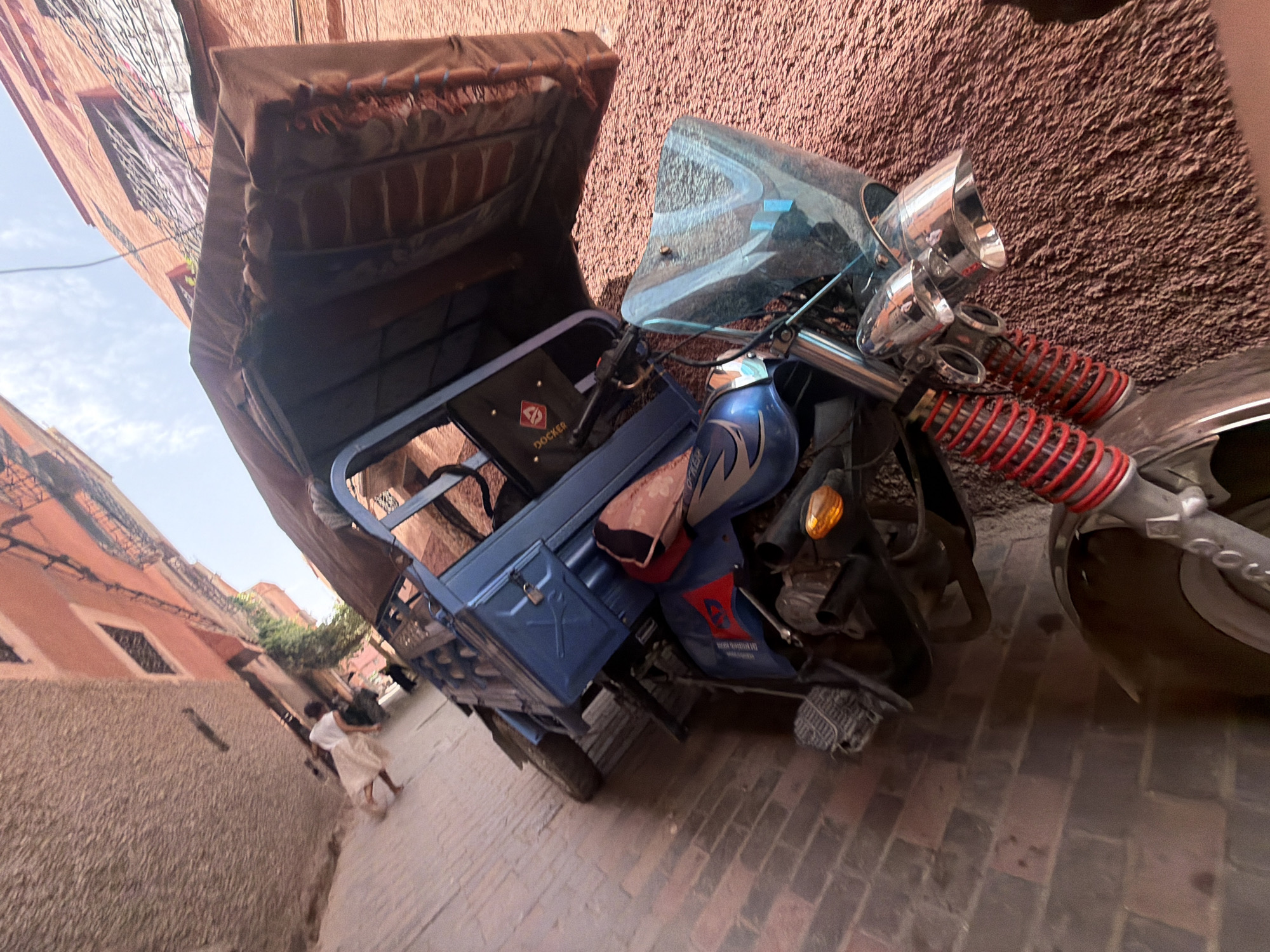 The image shows a street scene in Marrakesh, Morocco, likely in the medina given the narrow alleyway and traditional architecture. A blue three-wheeled auto rickshaw, commonly used for transport in the city, is parked on a brick-paved street. The auto rickshaw has a tan canopy and a blue cargo box. The buildings lining the alleyway are made of reddish-pink brick and have traditional windows with decorative grilles. A person, possibly a tourist, dressed in a white top and a light-colored skirt, walks away from the camera in the background. The lighting suggests it is daytime, with a bright sky visible between the buildings. The overall scene depicts a typical street view of Marrakesh, highlighting its unique urban landscape and modes of transportation.