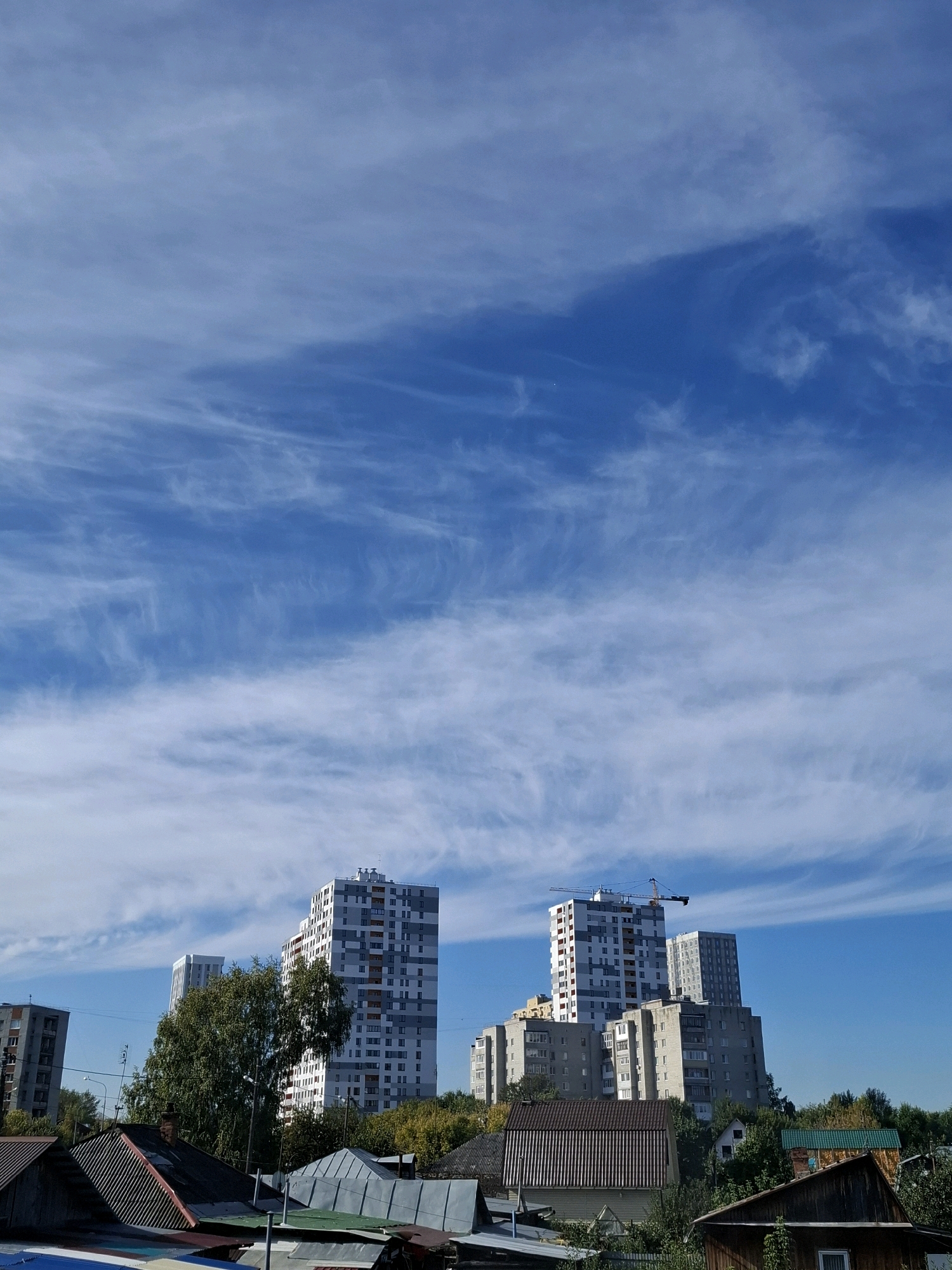 This image depicts a residential area in Tyumen, Russia, on a clear day. The sky is a vibrant blue, with wispy white cirrus clouds streaking across the upper portion of the frame. In the middle ground, several multi-story apartment buildings are visible, some with modern architecture featuring balconies and numerous windows. One building is under construction, indicated by a crane. In the foreground, a cluster of older, low-rise buildings with corrugated metal roofs and chimneys are present, suggesting a mix of older and newer construction within the neighborhood. Dense green trees are interspersed among the buildings, and a street lamp is visible on the left side. The overall scene conveys a typical urban landscape with a blend of residential structures.