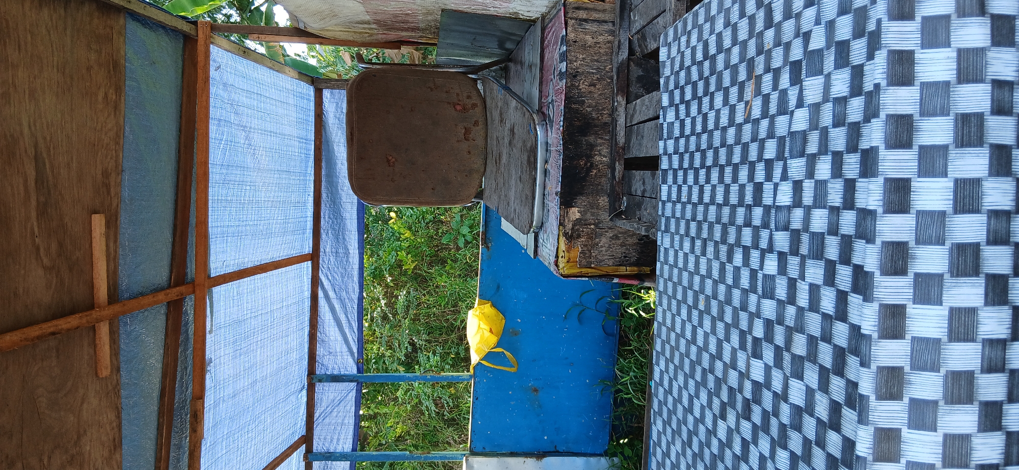 The image depicts an outdoor seating area, likely in a rural or semi-rural setting in Samarinda, Indonesia. In the foreground, a table is covered with a black and white checkered woven plastic mat. Behind the table, there is a raised platform made of wood and what appears to be a wooden crate covered with some form of textured material. On this platform sits an old, brown, upholstered chair with a worn seat and back.
To the left of the chair, a bright yellow plastic bag hangs on a blue metal barrier. Beyond the blue barrier, lush green vegetation and trees are visible, suggesting an outdoor environment. The structure overhead is covered with a blue tarp supported by wooden beams. The overall impression is one of a simple, functional, and perhaps makeshift outdoor seating area, possibly part of a food stall or a resting spot in a garden or roadside location within Samarinda.