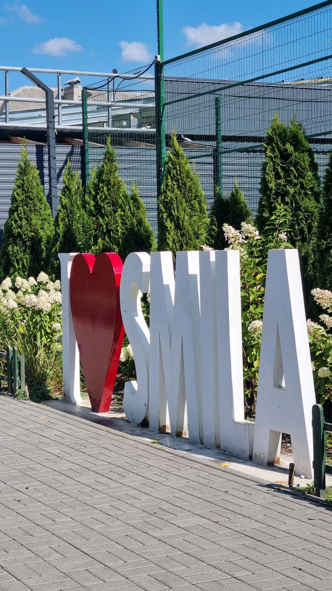 An outdoor, eye-level, full shot captures a large, three-dimensional "I Love Smila" sign made of white letters and a red heart. The sign is situated on a paved walkway made of gray rectangular bricks. Behind the sign, a line of evergreen trees forms a backdrop, interspersed with white hydrangeas. Further behind the trees, a dark gray corrugated metal wall is visible, with a wire mesh fence in front of it and a blue sky with scattered white clouds above. The setting appears to be an outdoor recreational area or public space in the city of Smila, Ukraine, given the signage.