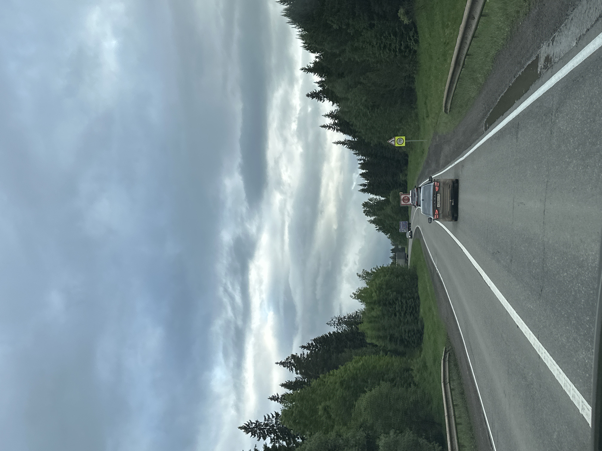 The image shows a road in Meshcheryaki, Russia. Cars are traveling on the asphalt road, which has a white dividing line. A yellow speed limit sign is visible on the side of the road, along with green trees and a metal guardrail. The sky is overcast with gray and white clouds.