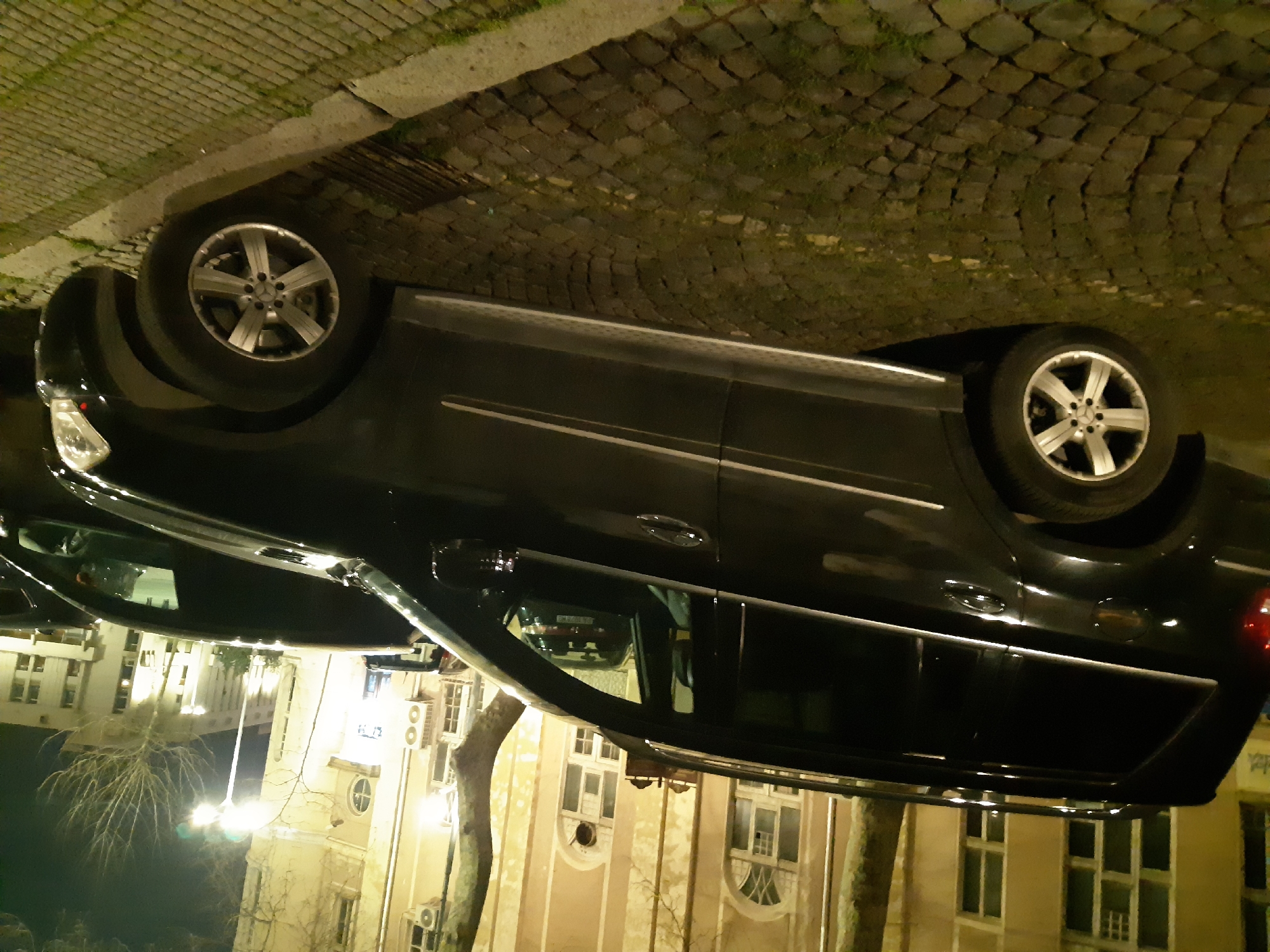 A dark-colored car is parked in Plovdiv, Bulgaria, alongside a cobbled street adjacent to a light-colored building with multiple windows. The car has silver rims on its tires, and its reflection is visible in the car's surface, showing more of the building. A bare tree branch is in the foreground.