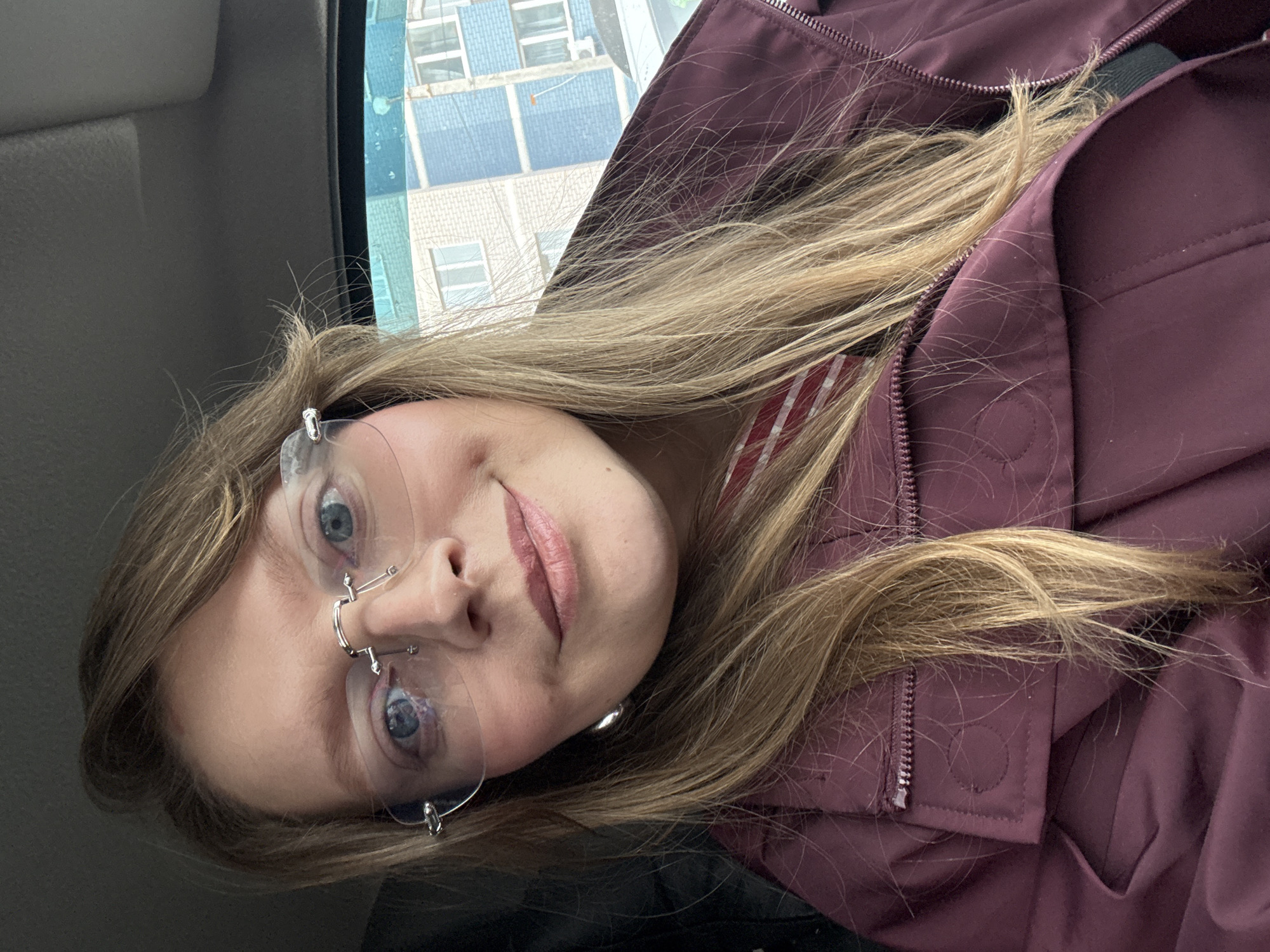 A woman with blonde hair and rimless glasses is angled facing the camera. She is wearing a burgundy jacket. Through the window behind her, a building with blue and white windows is visible. The location may be Izhevsk, Russia.