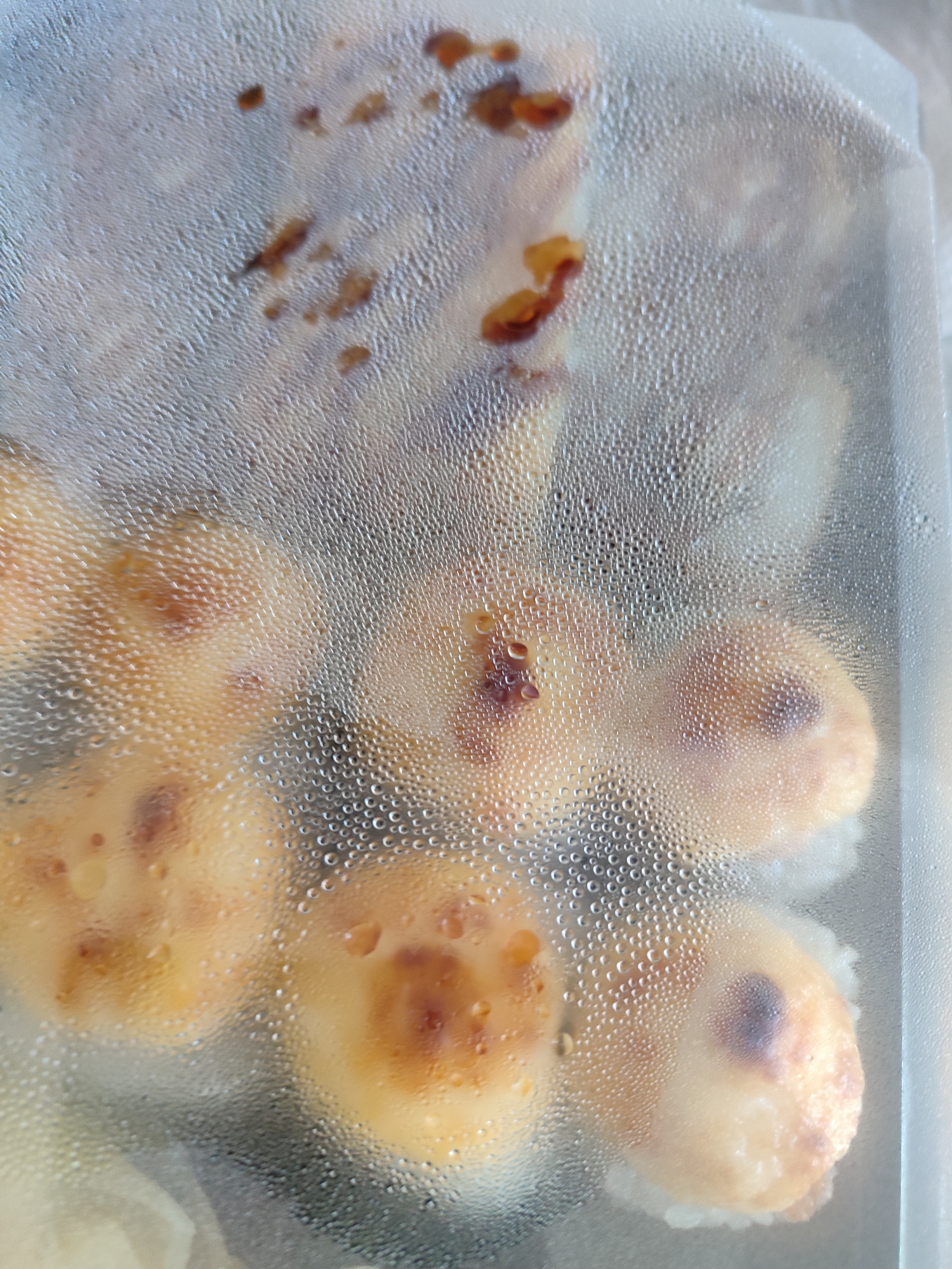 The image shows a close-up, overhead view of several pieces of sushi inside a clear plastic container. The lid of the container is covered in condensation, creating a blurry, textured effect over the food. The sushi appears to be baked or seared, with a golden-brown topping that has some darker, charred spots. The rice base of the sushi is visible underneath the topping. The lighting is diffused, and the overall impression is of a prepared food item ready for consumption, possibly taken from a takeout order. There is no visible text in the image. The context of Sochi, Russia, is not directly evident from the visual information alone.