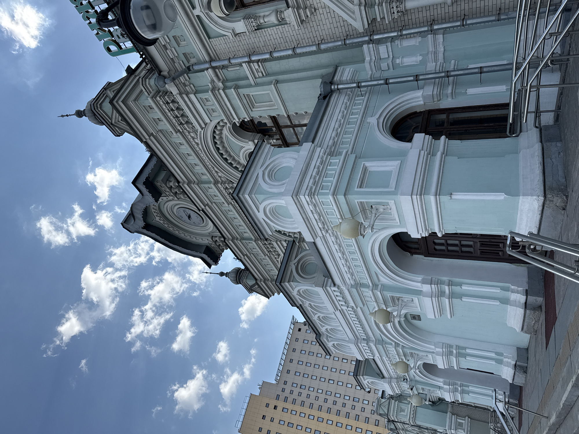 The image shows the facade of the Belorussky railway station in Moscow, Russia. The building is a pale blue color with ornate white trim and features arched windows and doorways. A clock tower is visible on the upper left, and a modern high-rise building is seen in the background. The sky is blue with scattered white clouds. Metal railings are present in the foreground.
