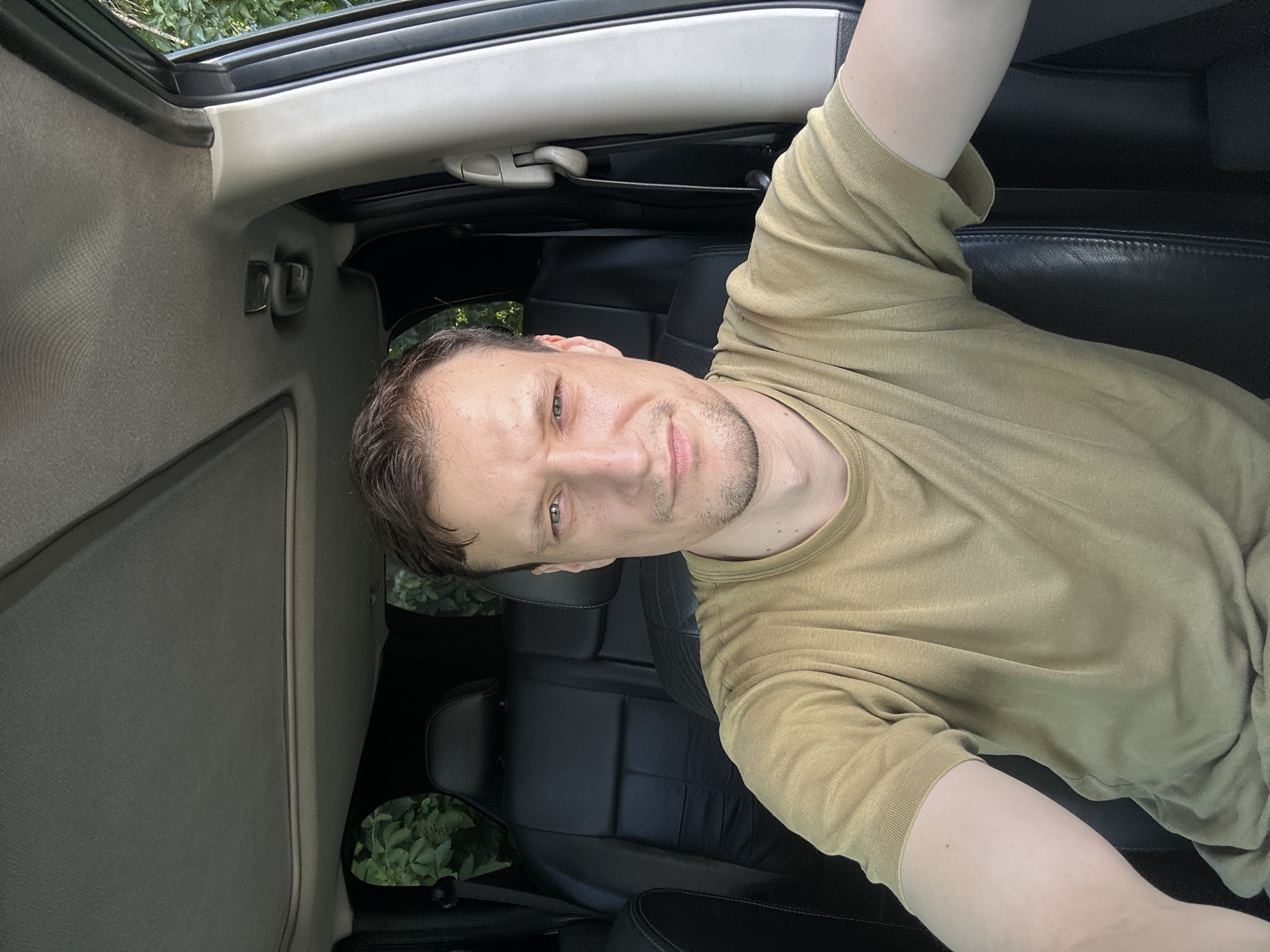 A man with short brown hair and a light beard is sitting in the driver's seat of a car, looking directly at the camera. He is wearing a short-sleeved olive green t-shirt. The interior of the car is visible, including black leather seats and a sunroof. Outside the car, green foliage can be seen. The image appears to have been taken outdoors during daylight. There is no specific indication of location within the city of Shatava, Ukraine, in the image itself.