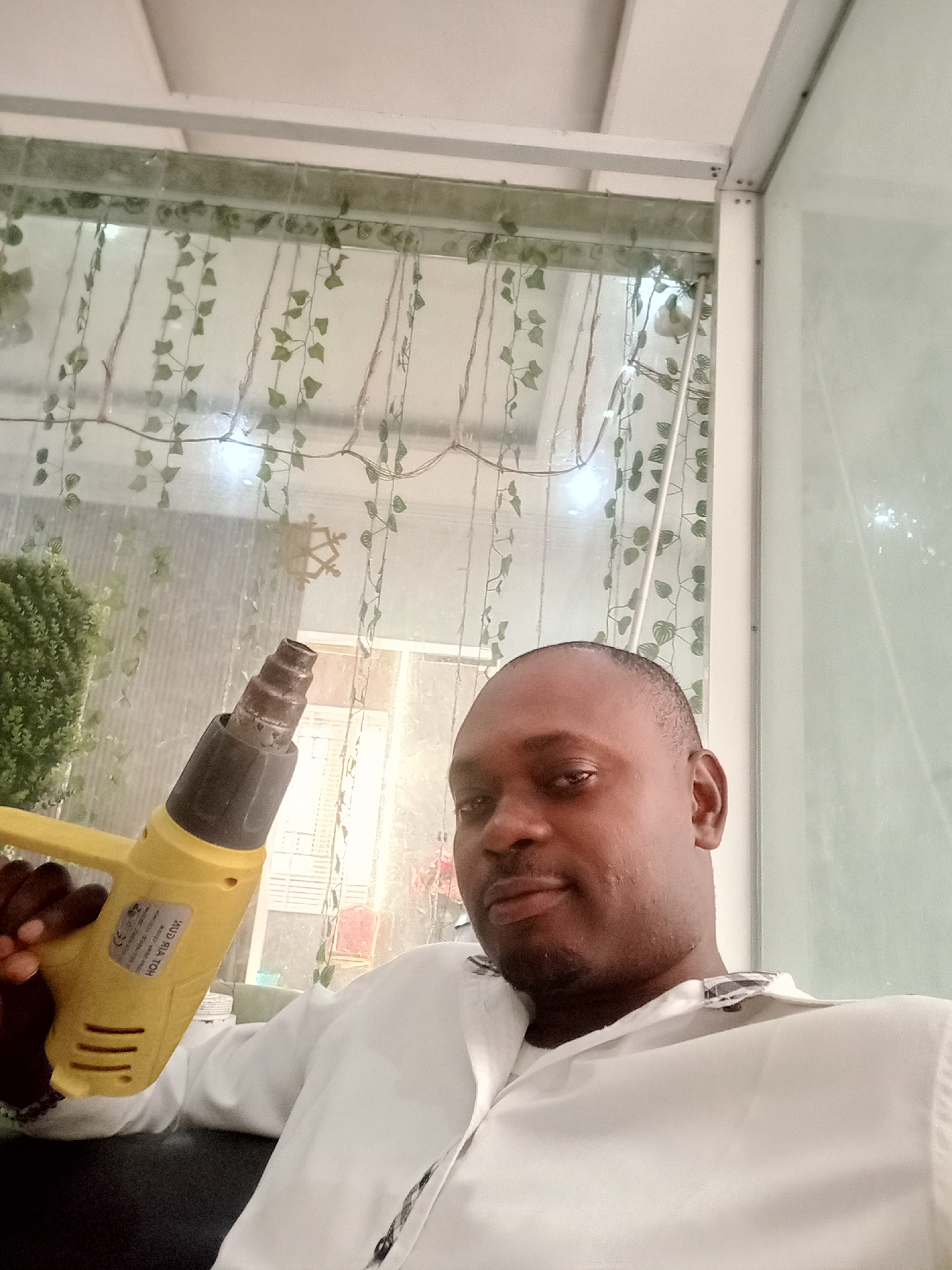 A man is holding a yellow heat gun in what appears to be an indoor setting. The man has dark skin and is wearing a white shirt. The background features a window with hanging green decorations. Based on the prompt's specifications, this scene is located in Gwarinpa, Nigeria.