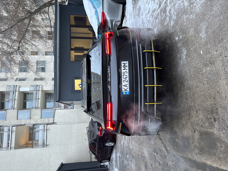 A black Lamborghini Urus, parked outside a building, exhibits its rear design. The car's license plate reads "KA 2475 MM." Striking yellow accents highlight the rear bumper. Exhaust fumes emanate from the vehicle, hinting that it may be running or recently turned off. The backdrop includes an apartment building and a glimpse of bare trees.