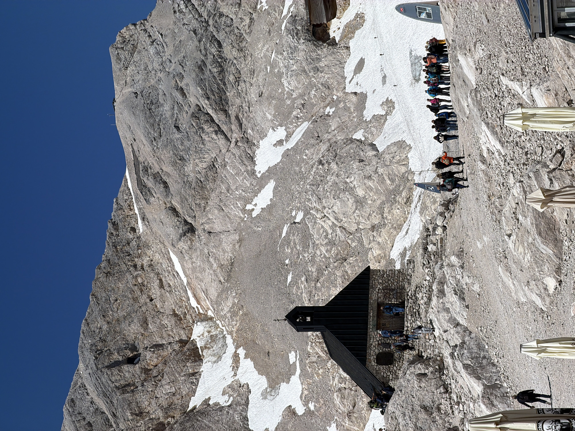 The image shows a mountainous landscape with a clear blue sky. In the foreground, there is a small, dark-colored chapel with a stone base. Several people are gathered around the chapel and on the rocky terrain leading up to it. A group of people stands further back, looking towards the chapel or the surrounding mountains. In the distance, snow patches are visible on the rocky slopes of the mountain. Partially visible in the lower foreground are several closed outdoor umbrellas. The scene appears to be a tourist destination, likely a mountain summit or viewpoint. This location is the Zugspitze, the highest peak in Germany, near Garmisch-Partenkirchen.