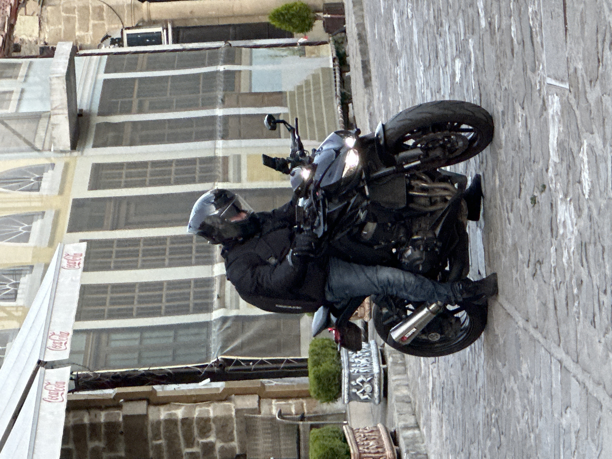 A person wearing a black helmet, black jacket, and blue jeans is sitting on a black motorcycle. The motorcycle has its headlights on and is parked on a cobblestone street. In the background, there is a building with awnings that have the Coca-Cola logo. The building has many windows and a stone facade. To the left of the building, there is a stone wall. To the right of the building, there is a small, potted evergreen tree. The scene appears to be in a city.