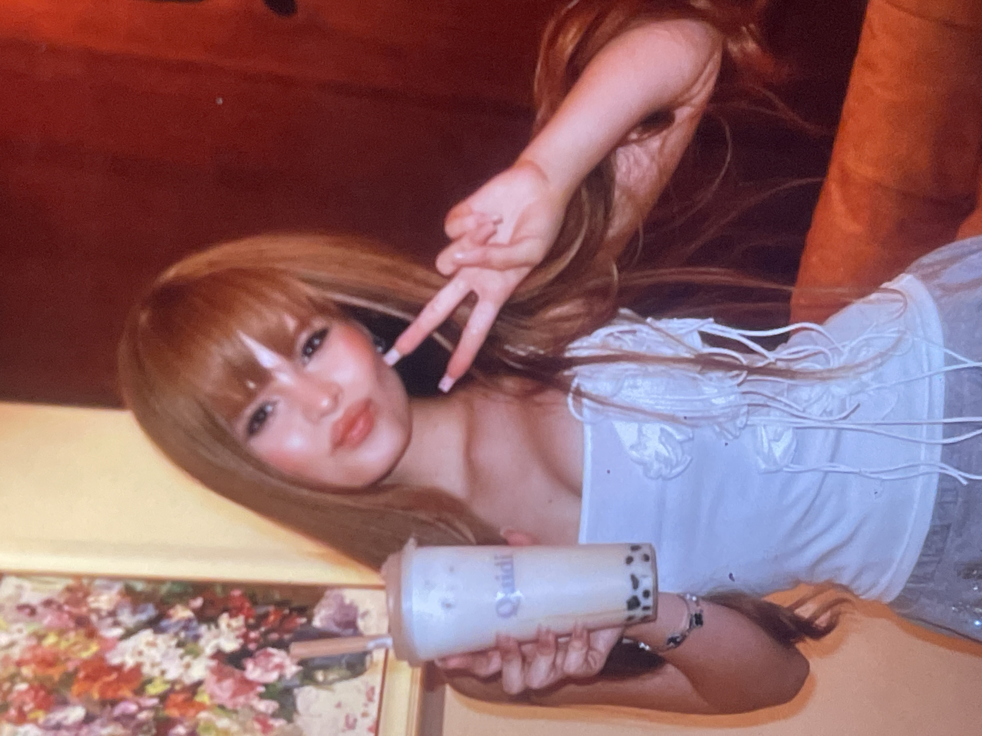 A young woman with long, light brown hair and bangs is depicted in a close-up, rotated portrait. She has light makeup, including winged eyeliner and rosy blush, with a soft smile. Her left hand is raised, making a peace sign, while her right hand holds a tall, clear cup of boba tea. The drink inside is light-colored, possibly milky, with dark boba pearls visible at the bottom. The brand "Qaidi" is visible in purple text on the side of the cup.
She is wearing a white top with intricate ruffled details and dangling white strings on the front. The background on the upper left side is a warm, reddish-brown, possibly a wall or curtain, while the lower right reveals a lighter, possibly yellowish, surface. In the bottom left corner, part of a framed artwork, resembling a floral painting with vibrant red, pink, and green hues, is visible.
The activity appears to be posing for a casual photograph indoors, likely in a cafe or a well-lit room with warm ambient lighting. The atmosphere seems relaxed, and her expression suggests contentment. No specific location cues are visible within the image itself to confirm the provided context of Poltava, Ukraine.