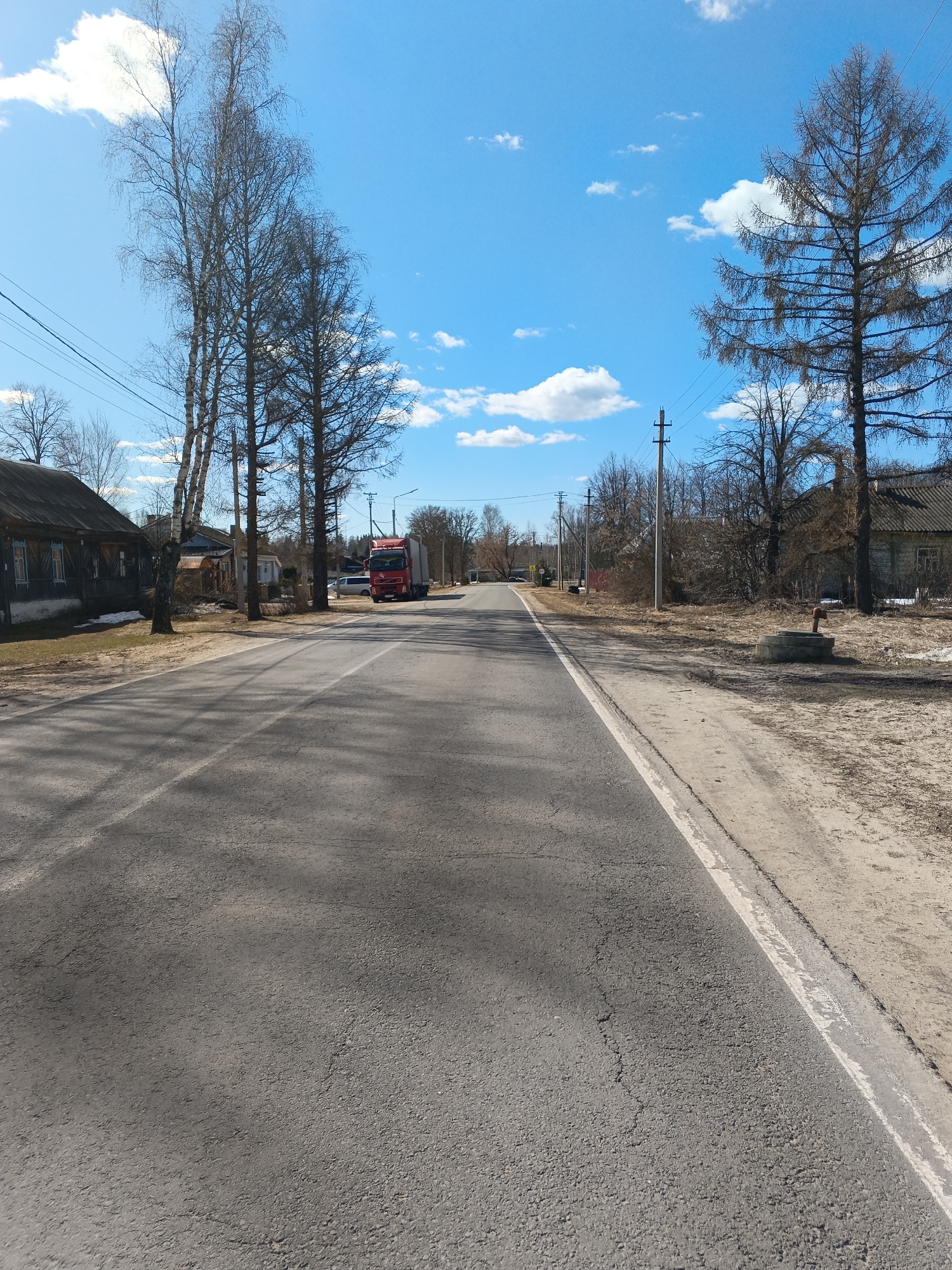 The image depicts a straight, two-lane asphalt road in Красный Маяк, Russia. The road stretches into the distance with a white line marking the right edge. On either side of the road are dirt shoulders bordering rural landscapes. The sky is blue with scattered white clouds. Along the road, there are utility poles, bare trees without foliage, and modest houses with dark wooden exteriors. A large red semi-truck is parked on the left side of the road, with a smaller white vehicle visible behind it. A concrete well or water access point is located on the right side of the road. The scene conveys a rural or semi-rural setting, likely in early spring given the absence of leaves on the trees.