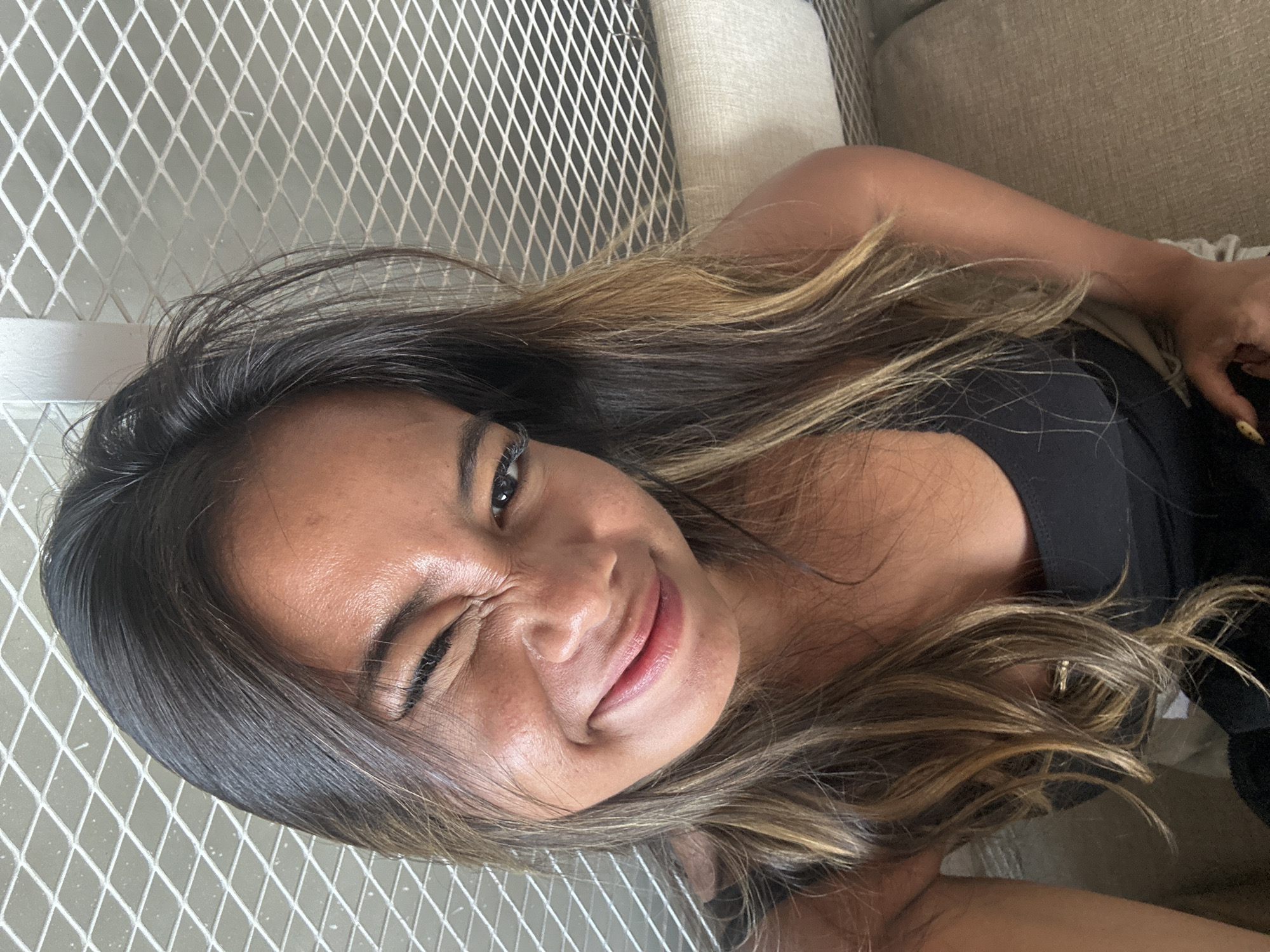 In North Kuta, Indonesia, a young woman is captured in a close-up selfie. Her hair is long and wavy, with highlights of blonde and brown, framing her face. She is winking with her right eye and smiling broadly, her lips slightly parted, revealing her teeth. Her skin appears sun-kissed and slightly shiny. She is wearing a black tank top. The background consists of a diamond-patterned metal mesh structure and a portion of a textured beige couch. The lighting suggests an indoor setting, possibly during the daytime.