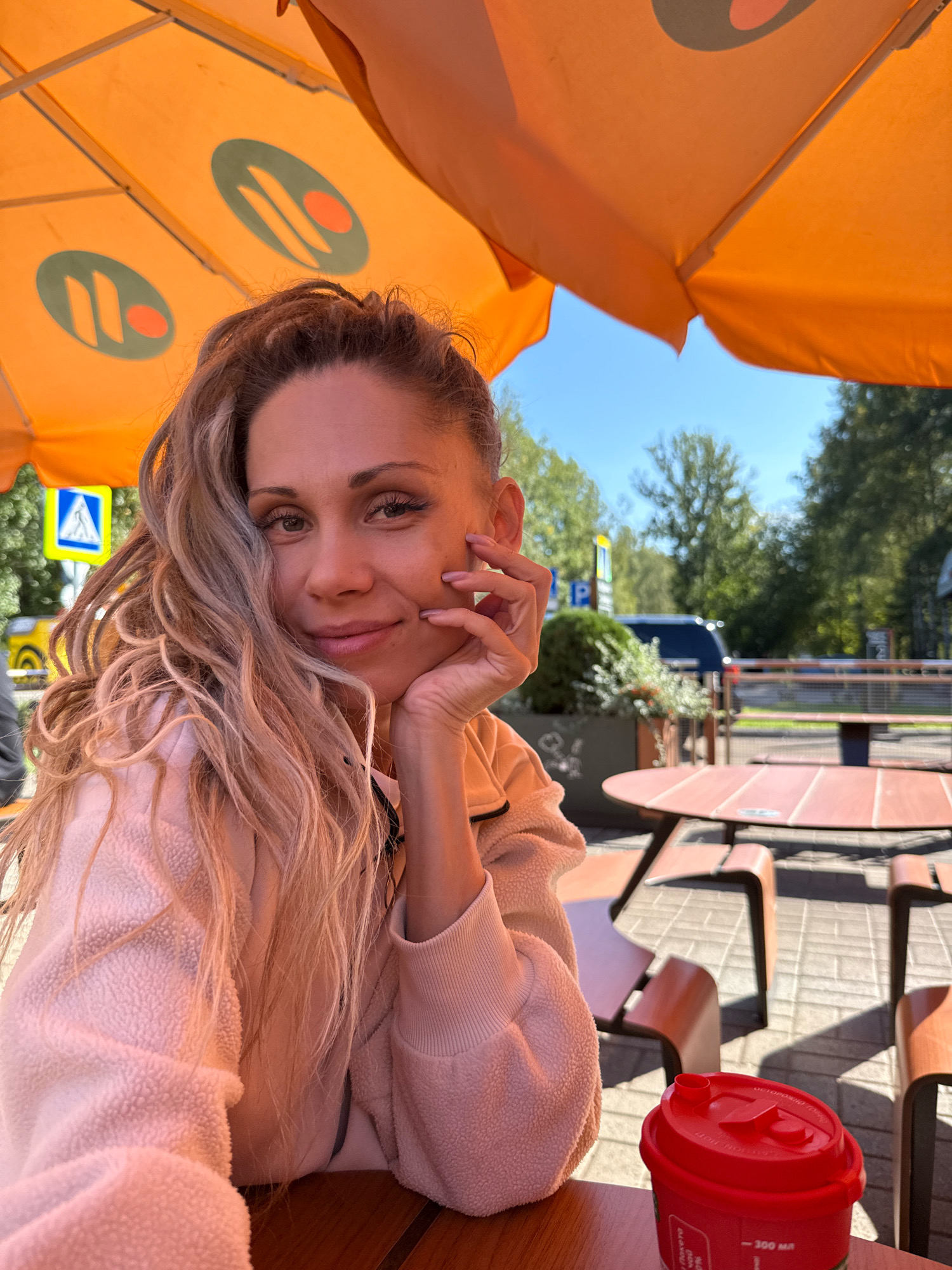 A woman with blonde hair is sitting at a table under an orange umbrella with a logo of two green ovals with orange shapes inside. She is wearing a light pink fuzzy jacket and resting her chin on her hand, looking at the camera. A red cup is on the table in front of her. The background shows a street with trees, a blue car, and a parking sign. This appears to be an outdoor seating area, possibly at a cafe or restaurant, in the city of Zhukovo, Russia.