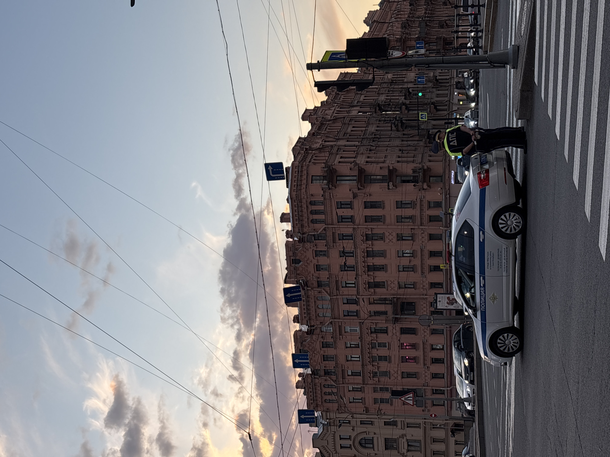 An eye-level street scene in Saint Petersburg, Russia, is captured at dusk, characterized by the soft, warm hues of sunset illuminating the sky. In the foreground, a uniformed police officer, wearing a high-visibility vest with "ДПС" (likely ДПС, or Road Patrol Service) emblazoned on it, stands beside a white police car. The car features blue stripes and prominent Cyrillic text "ПОЛИЦИЯ" (POLICE), along with contact information: "ТЕЛЕФОН ДОВЕРИЯ" (TRUST PHONE) and "ТЕЛЕФОН ДЕЖУРНОЙ ЧАСТИ" (DUTY UNIT PHONE) with the 8 (812) area code. The officer and vehicle are positioned near a pedestrian crossing.
Dominating the background is a large, ornate, multi-story building with reddish-brown/pinkish facades, indicative of classic European architecture common in Saint Petersburg. Overhead, a complex network of power lines crisscrosses against the pastel-colored sky with scattered clouds. Various street signs, including blue directional arrows and traffic signals, are visible throughout the frame, reinforcing the urban setting. A partially visible sign with the text "аптека" (pharmacy) can also be seen. The scene depicts a routine evening in the city.