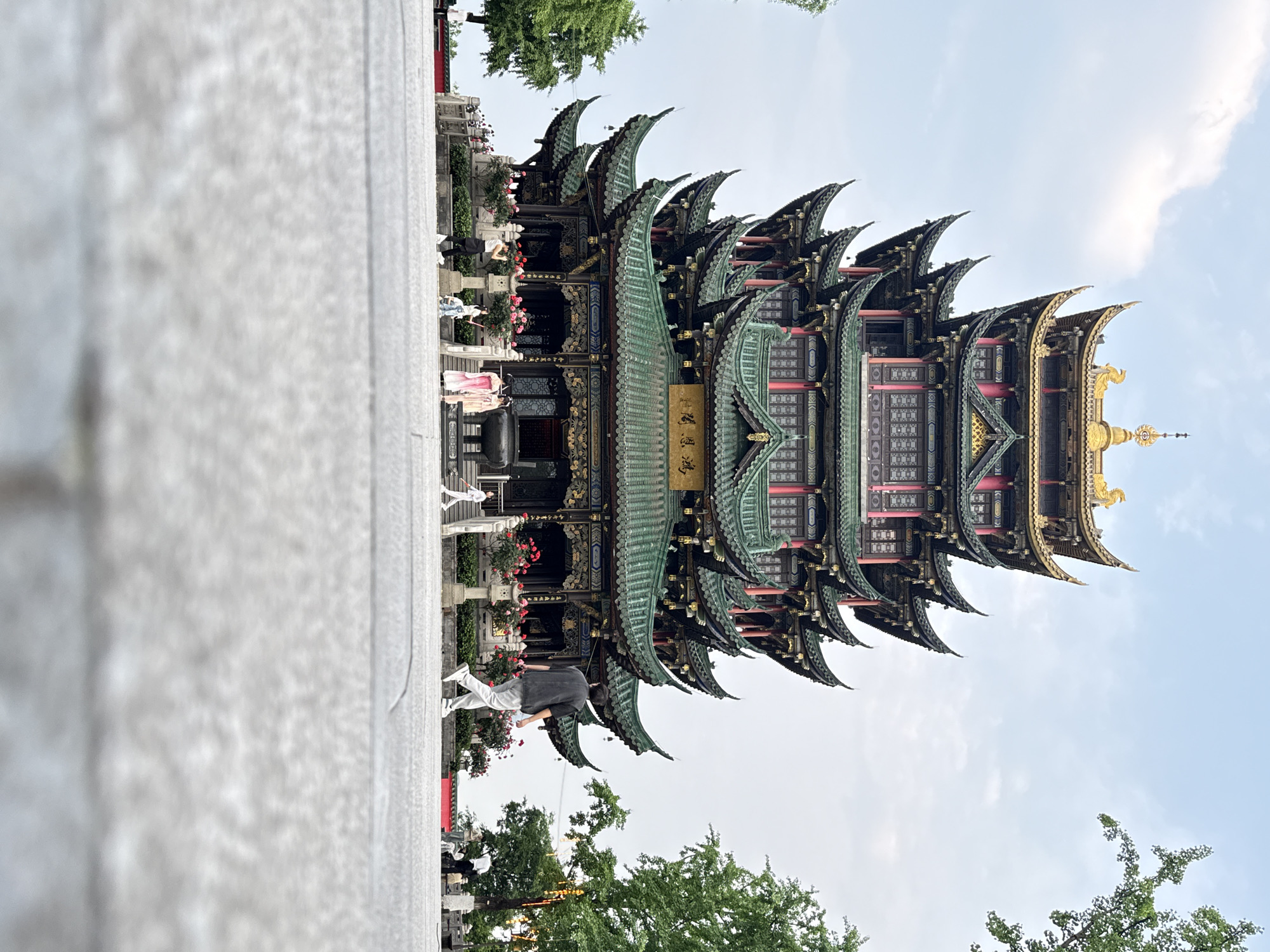 The image depicts a towering, multi-tiered pagoda structure, likely a landmark, against a backdrop of a light blue sky with scattered clouds. The architecture is distinctly Chinese, with upturned eaves and intricate details in green, gold, and red. People are seen near the base of the structure; one is dressed in a light-colored traditional gown and another is wearing a dark grey t-shirt with white pants, the latter appears to be walking. Lush greenery and trees frame the bottom of the image. The foreground is dominated by a large concrete structure. Based on the mention of Liangjiang, China, the location of the pagoda is most likely somewhere in the Liangjiang New Area of Chongqing.