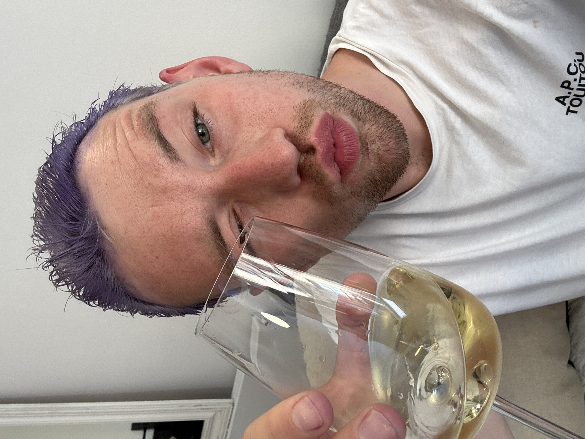 A close-up, angled portrait captures a man with violet hair holding a wine glass filled with a pale yellow liquid. The man's face is close to the camera, showing light stubble, prominent lips, and an eye looking toward the viewer. He wears a white t-shirt with the letters "A.P.C. TOUITOU" printed on it. The background includes part of a white wall, a gray or beige couch, and a white-framed mirror. The location is not discernible from the given image.