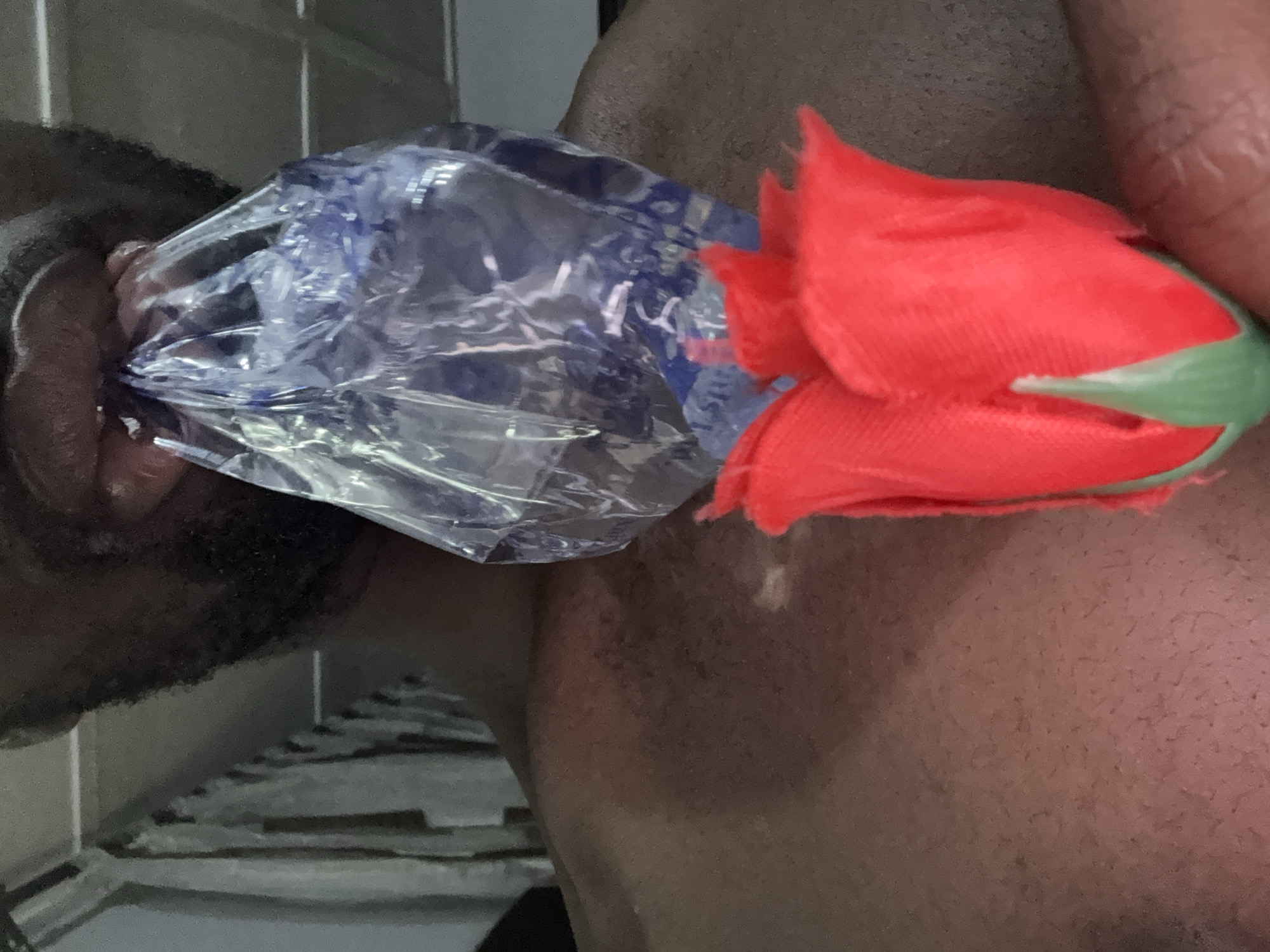 A close-up shot captures the upper torso of an individual with a dark complexion and a beard. The individual's mouth is open, gripping a crumpled, translucent plastic bag that appears to contain a blue substance or liquid. A single artificial red rose, with a green stem base and sepals, is positioned in the lower-right foreground, extending upwards towards the individual's chest. The rose's petals exhibit a textured, possibly fabric-like, appearance. A hand is partially visible near the base of the rose, with fingers lightly touching it. The individual's chest and shoulder area are exposed, suggesting they are shirtless or wearing dark, indistinct clothing. The background is dimly lit and out of focus, revealing vertical and horizontal structures consistent with an indoor setting, possibly shelving units, a clothes rack, or blinds. The location is an unknown, unknown city.