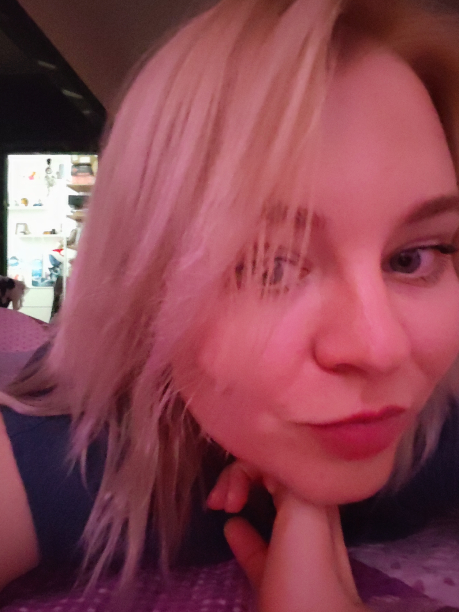 A close-up image features a woman with blond hair. She is looking towards the camera. Her left hand is positioned under her chin. There's a hint of furniture and a shelf in the background. The lighting appears to be red. There is no evidence in the image to suggest that the image was taken in Izhevsk, Russia.