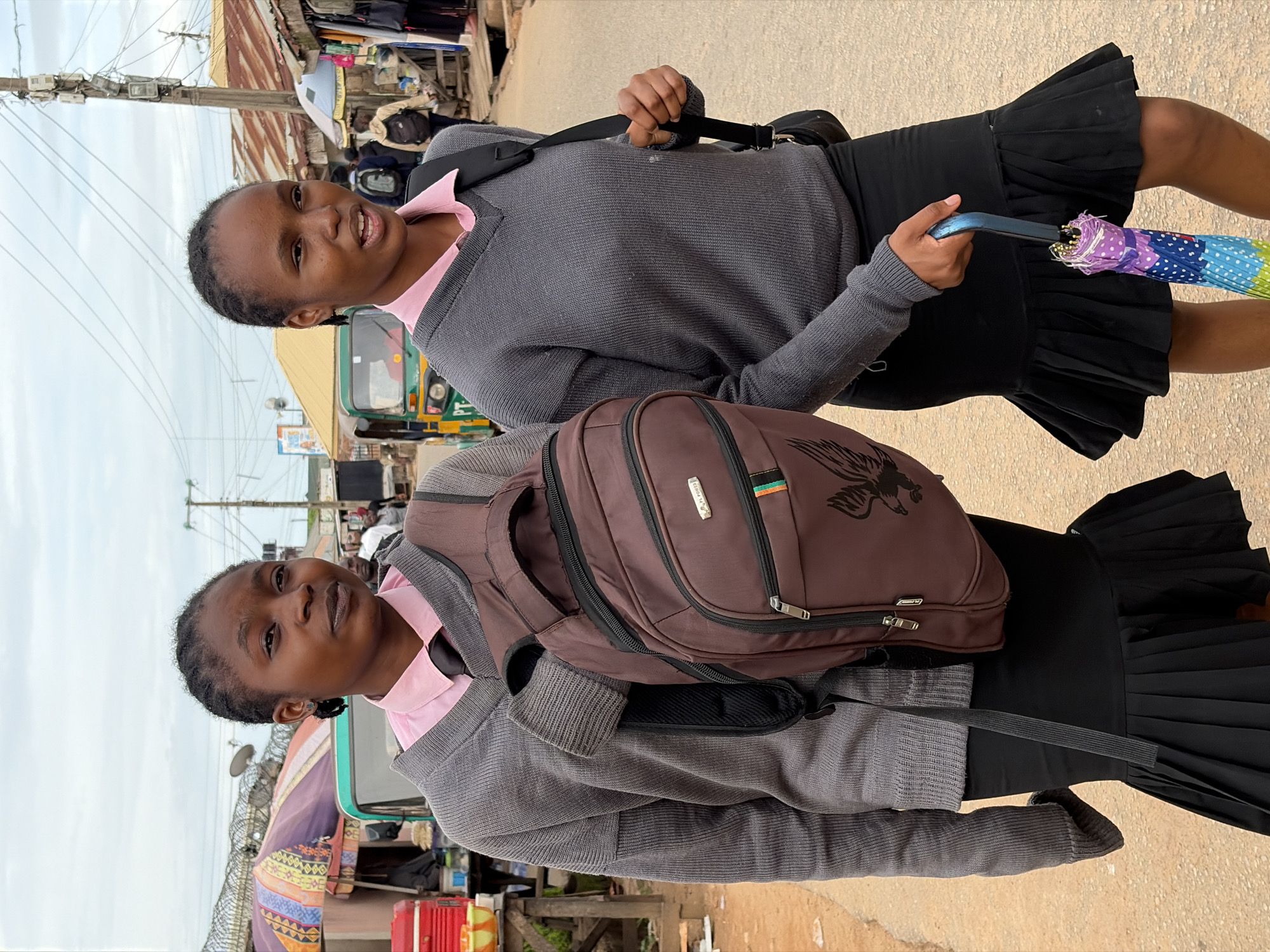 Two young women, presumably students, are walking on a street in Jos, Nigeria. The woman on the left has her hair in braids and is wearing a grey V-neck sweater over a pink collared shirt and a black tie. She is carrying a brown backpack. The woman on the right is also wearing a grey V-neck sweater over a pink collared shirt and a black tie. She has a black strap from a bag over her shoulder and is holding a colorful umbrella. The street appears to be a market area with stalls and vehicles, including a three-wheeled auto-rickshaw. Overhead power lines are visible, along with utility poles. The ground is a dirt or gravel road.