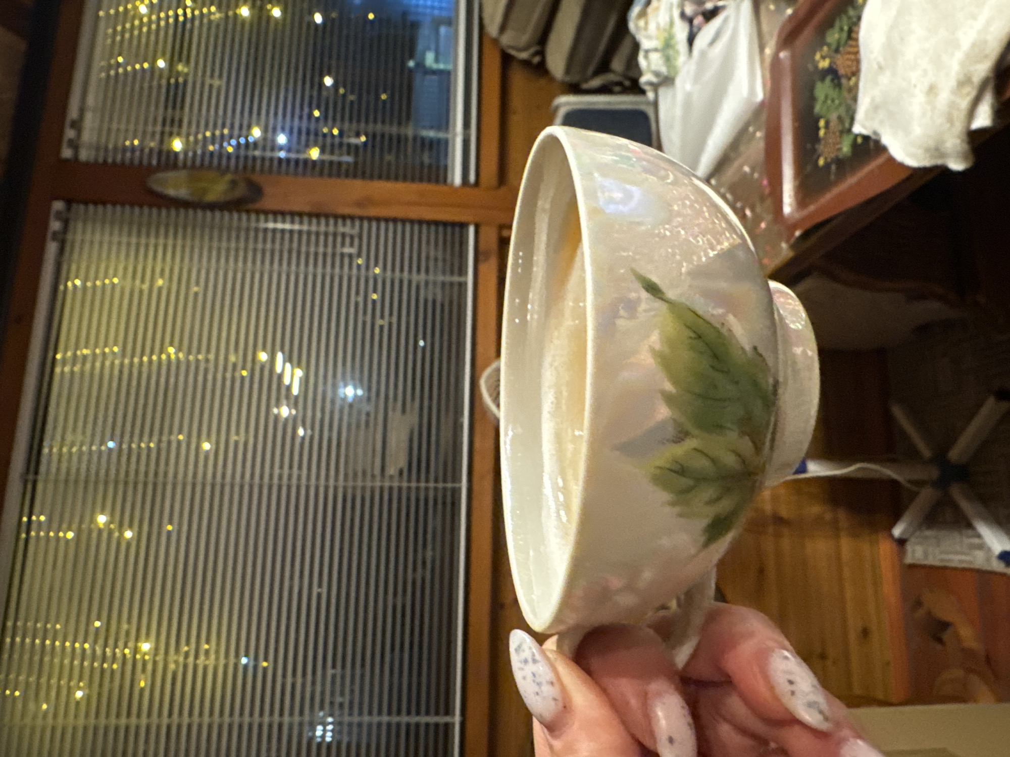 A person holds a teacup in front of a window. The teacup is white with a pearlescent sheen and a green leaf design. The window has vertical blinds, and outside, there are lights, likely from a city at night. Other objects are in the background but are blurry, including items on a shelf. The person's hand is visible with long, decorated nails. The location may be in Красный Маяк, Russia.