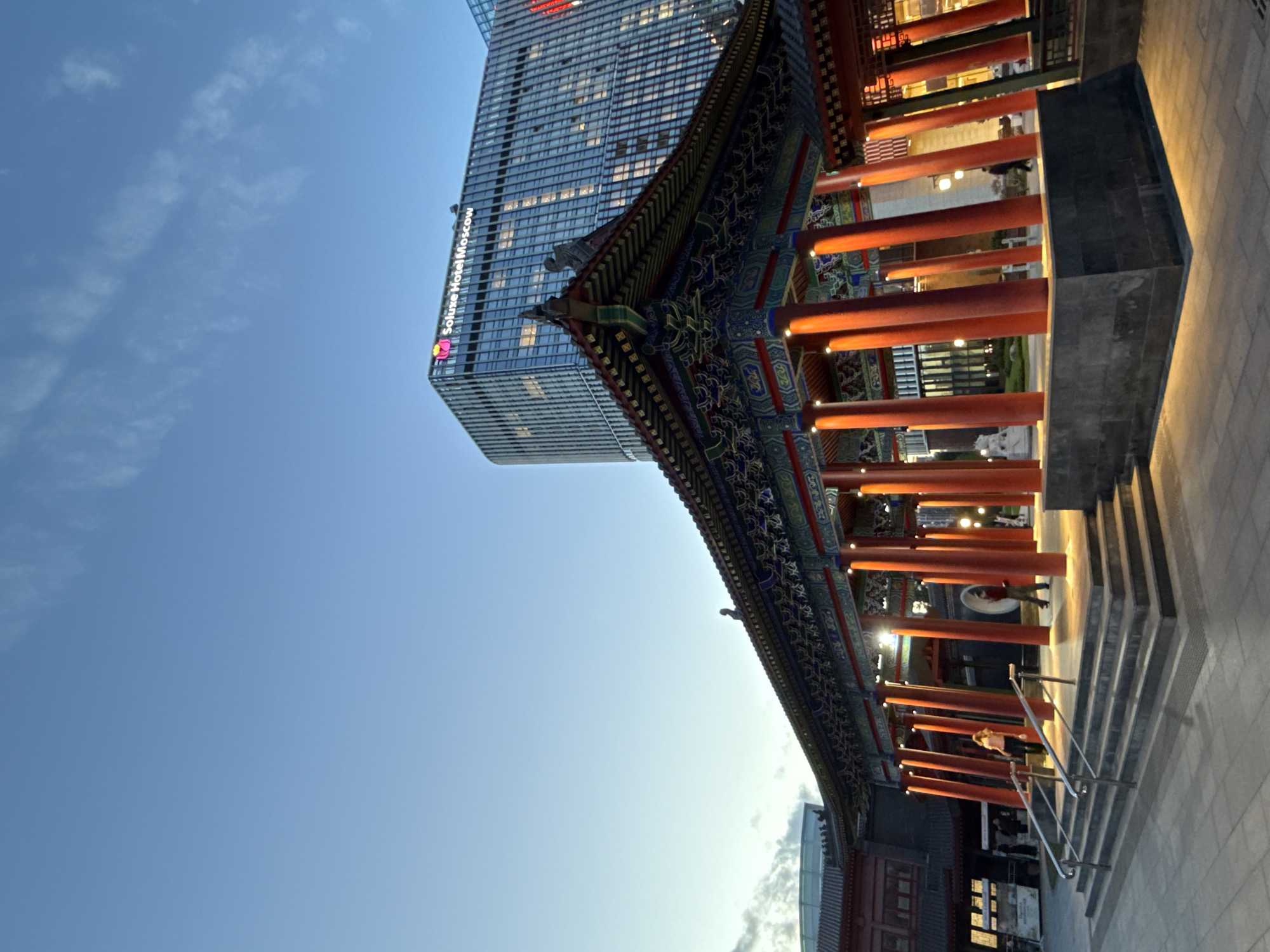 In Moscow, Russia, a traditional Chinese pavilion with red pillars and ornate eaves stands in the foreground, illuminated by warm artificial lights. The pavilion is situated on a paved plaza with wide steps and handrails leading up to it. In the background, a modern skyscraper with the prominent signage "Soluxe Hotel Moscow" rises into the clear blue sky. The sky is a pale blue with wispy clouds, indicating either early morning or late evening. Several people are visible in and around the pavilion, some walking, and others seemingly observing the architecture. The scene juxtaposes traditional East Asian design with contemporary urban architecture, suggesting a cultural or commercial district within the city.