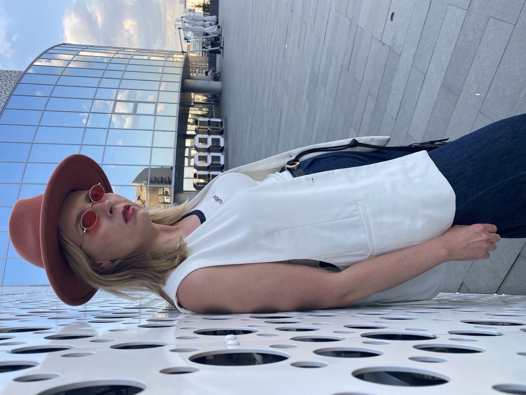 A woman wearing a brown hat and red sunglasses lies on a white perforated surface. She is also wearing a white vest and blue jeans. In the background is a modern building with a curved glass facade, reflecting the sky. Further back, people are visible on a terrace-like structure. The scene appears to be in an urban area, potentially Yekaterinburg, Russia, judging by the style of architecture and the urban setting.