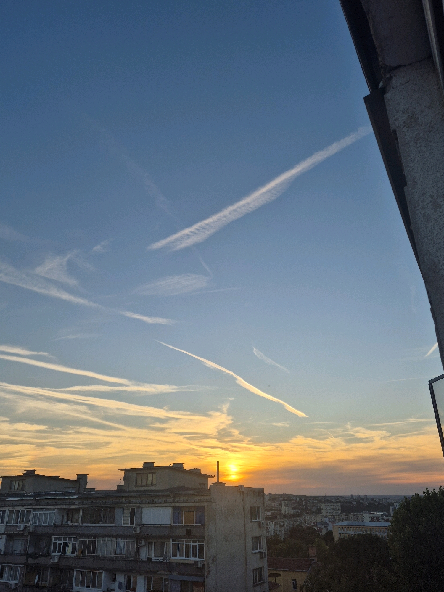 The image depicts a city skyline at sunset, with a dramatic sky filled with numerous contrails from airplanes. The sun is low on the horizon, casting a warm, orange glow across the clouds and the cityscape. In the foreground, a multi-story apartment building with numerous windows and balconies dominates the lower left and center of the frame. The building appears aged, with visible air conditioning units and architectural details typical of urban residential structures. Beyond the apartment building, a panorama of other city buildings and trees stretches into the distance, fading into the twilight. The sky above is a vibrant blue, transitioning to yellow and orange near the horizon. The right edge of the image is framed by a portion of a concrete structure and a dark, possibly metal, edge, suggesting the viewpoint is from a balcony or a rooftop. No people are visible in the image. The overall atmosphere is serene and evocative of the end of the day in an urban environment.