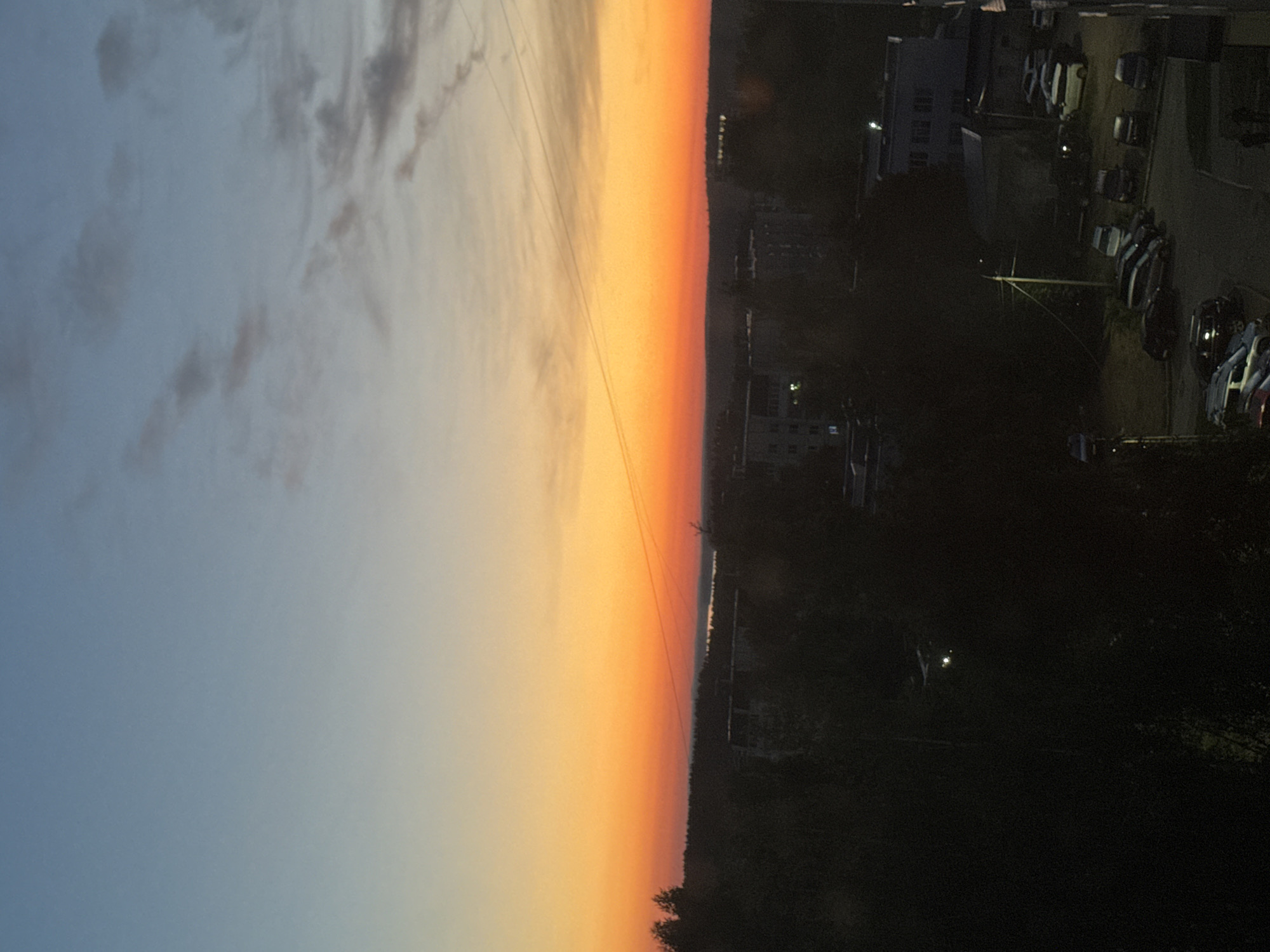 The image, when viewed horizontally, depicts a twilight scene, likely sunset, over an urban or suburban landscape. The sky dominates the upper and central portions, displaying a dramatic horizontal gradient. On the far left, the sky is a serene light blue, transitioning through areas with wispy grey clouds, into a vivid spectrum of orange and yellow hues as it approaches the horizon on the right. Delicate power lines or communication cables are visible stretching across the middle of the sky.
A clear horizon line separates the luminous sky from the dimly lit land below. Along this distant horizon, a dark silhouette of trees and scattered buildings is discernible. A faint, horizontal shimmer suggests the presence of a body of water or distant urban lights.
The foreground, which occupies the lower right portion of the image (when correctly oriented), is steeped in darkness, indicating nightfall. It reveals an urban environment featuring several dark building structures, some with visible windows or faint interior lights. A parking lot or street area is populated with numerous parked cars, identifiable by their shapes and occasional reflections. Scattered streetlights cast subtle glows, and the outlines of dark trees and bushes are visible.
The scene captures the transition from day to night, evoking a sense of quietude. The provided context indicates the location is Chaikovsky, Russia. There is no visible text in the image.
