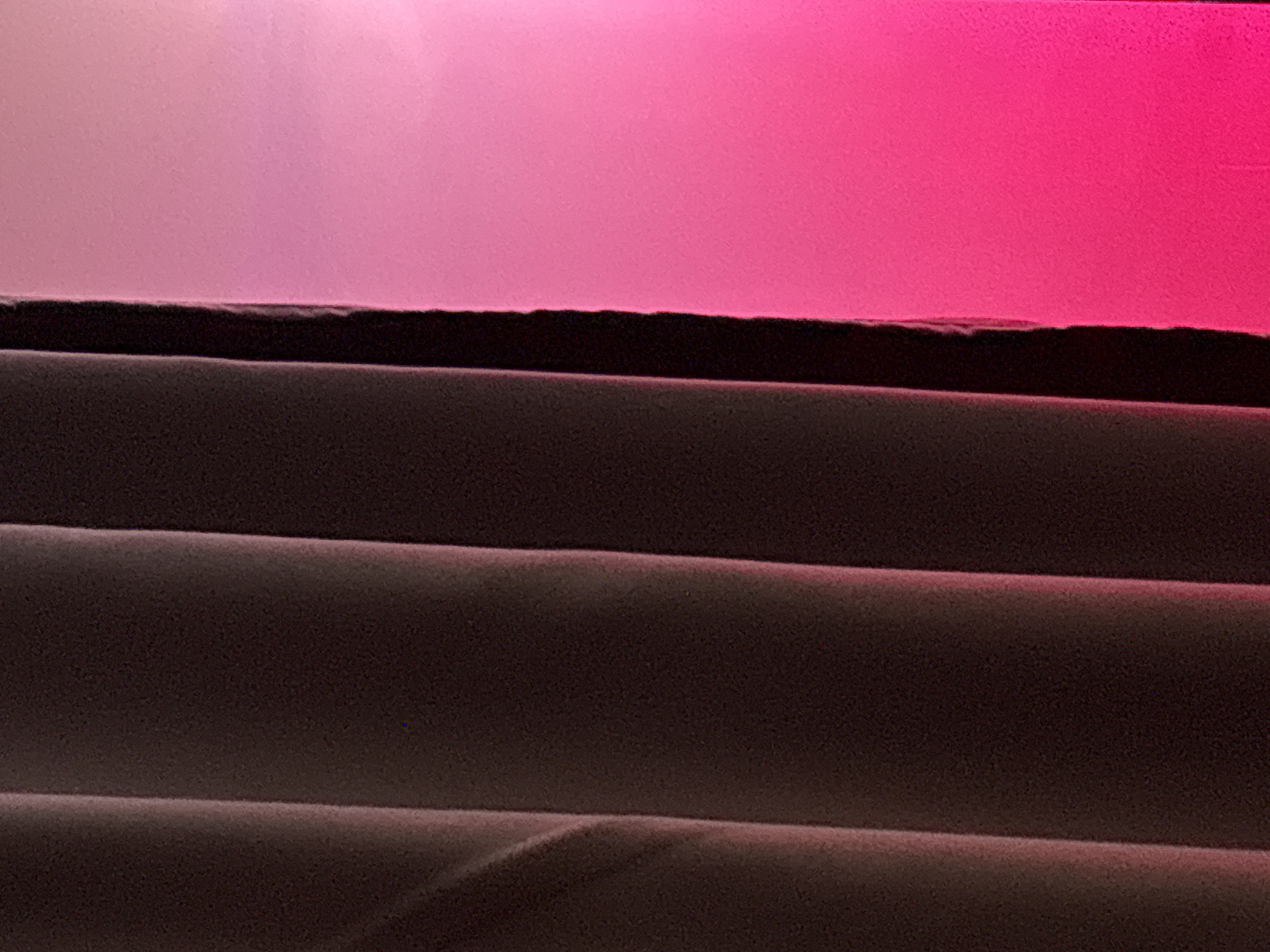The image depicts a close-up view of window blinds. The slats are dark brown and partially closed, creating horizontal lines. Above the blinds, a diffuse pink and white light is visible, suggesting an external light source filtering through. There is no indication of a location in Saint Petersburg or any other city, people, actions, or interactions in the image.