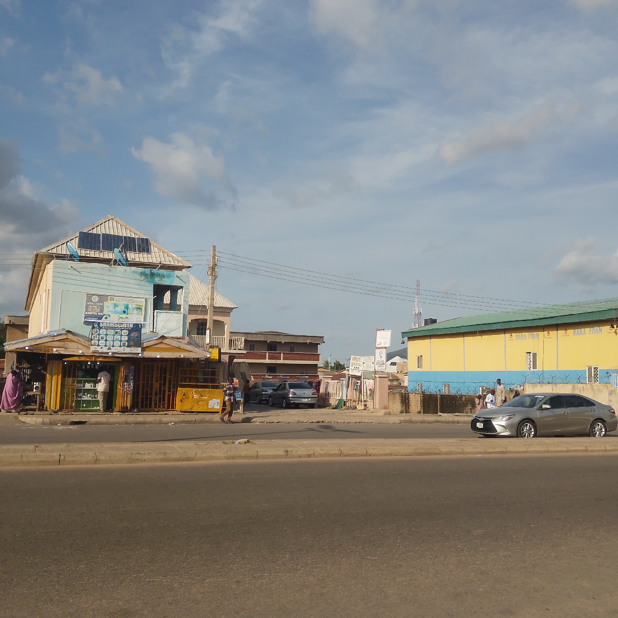 The image depicts a street scene in Pantame, Nigeria, under a partly cloudy sky. In the foreground, a paved road with a concrete divider separates the camera's perspective from the buildings and activity on the other side.
On the right side of the frame, a silver sedan is parked near the curb. Further down the street, a large, yellow and blue building with the words "HARA FOAM" is visible. Several people are standing in front of this building, some appearing to be customers or employees.
To the left of the Hara Foam building, other structures are present, including what appears to be a multi-story building with balconies and a smaller shopfront. In front of the shopfront, there are displays of goods. A person in a pink garment stands near the shop, and another individual in a white shirt is inside. A yellow stall is also visible to the left of the shop.
In the middle ground, several cars are parked on the side of the road. Behind the shopfronts, there are more buildings, some with multiple floors. A telephone pole with wires runs across the scene. The architecture suggests a mix of commercial and residential properties. The overall impression is of a busy commercial area in a Nigerian city.