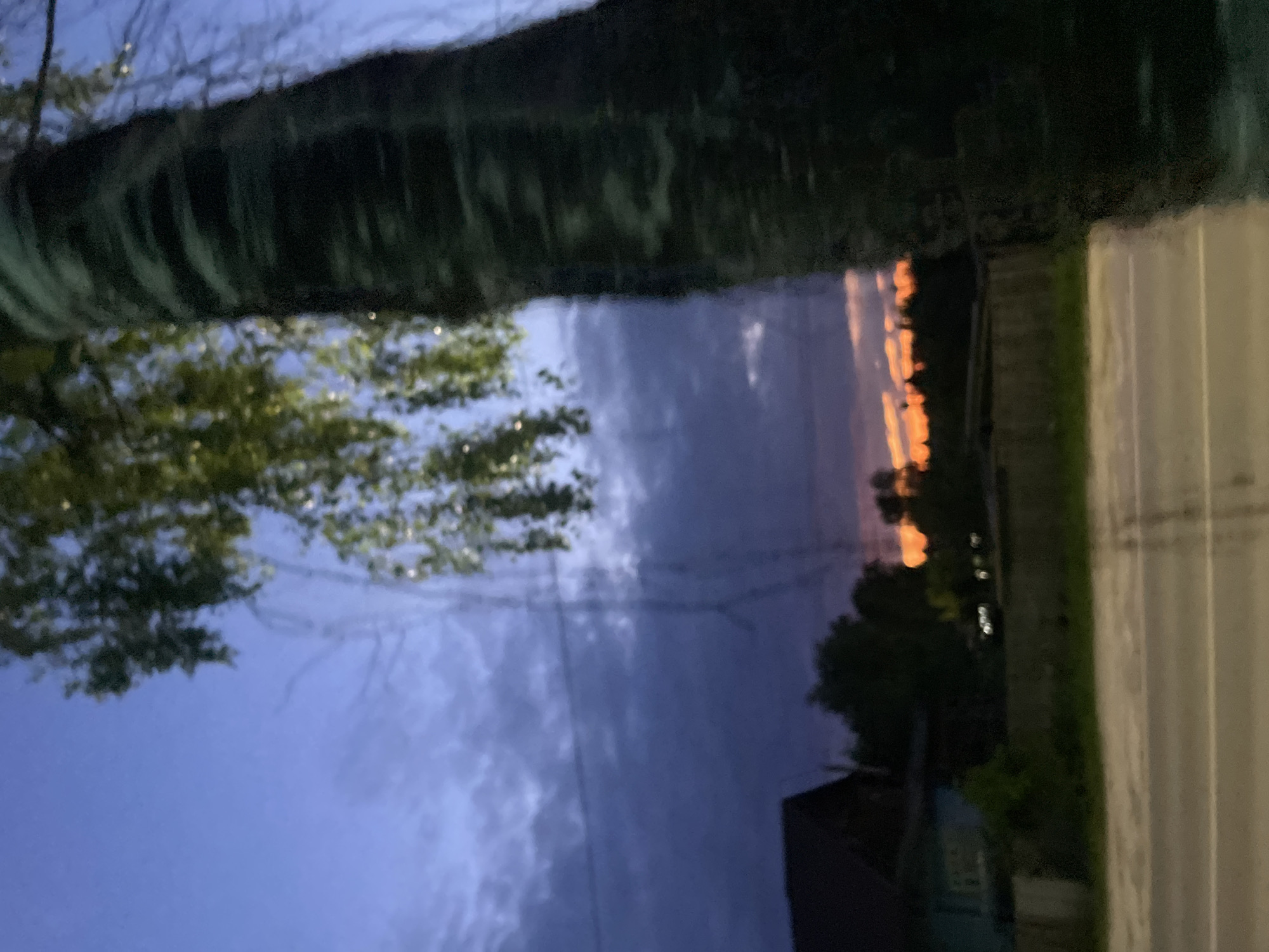 The image presents a rural or semi-rural landscape in Красные Баки, Russia, captured during dusk or dawn. A substantial, dark, and out-of-focus tree trunk occupies the right foreground, with blurry green foliage exhibiting sparse light reflections in the upper right. The sky, viewed through and beyond the tree, transitions from a muted blue-purple at the zenith, to a wide band of dark grey-purple clouds mid-frame, culminating in a prominent orange and red glow along the horizon. Silhouetted trees are discernible against the colorful sky. In the middle-ground on the left, a dark-roofed structure and a lighter-colored building are partially visible. Faint utility lines horizontally traverse the mid-left, intersecting with a translucent, vertical structure, possibly a utility pole or a reflection. The lower foreground consists of a paved road with white lane markings, flanked by a strip of green foliage and a low, dark fence or wall. No individuals are present, and no specific actions or interactions are depicted.