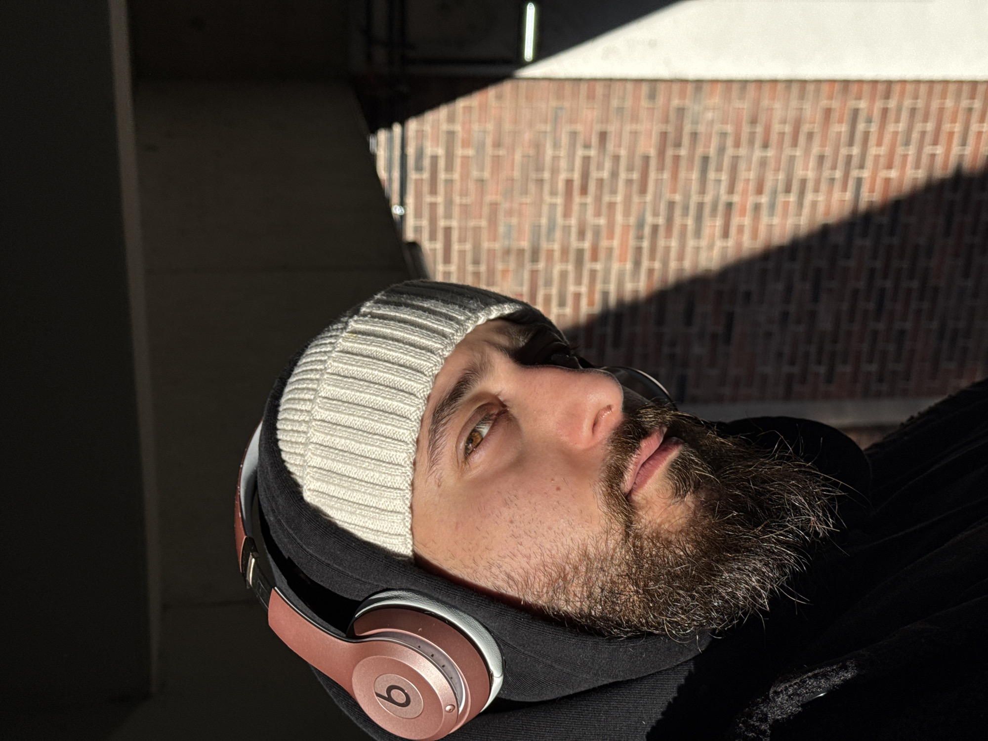 A male individual is depicted in a close-up, medium shot, looking directly at the viewer. He possesses a dark, full beard and mustache, and light brown eyes. He wears a light beige ribbed knit beanie topped with rose gold/copper Beats by Dre over-ear headphones. A black hooded garment is visible beneath the headphones and beanie, with a dark, possibly textured outer layer on his left side. Strong, directional sunlight illuminates the right side of his face, the headphones, and the background, casting significant shadows on the left side of his face and parts of the background. The immediate background on the right features a reddish-brown brick wall, partially lit by direct sun and partially obscured by a sharp, diagonal shadow. A brightly lit vertical strip of a light-colored wall or concrete structure is visible on the far right. Above the subject and the brick wall, a dark, shaded overhead structure is present, suggesting an underpass or building overhang. The scene is located in an unspecified urban environment within the city of Strasbourg, France.