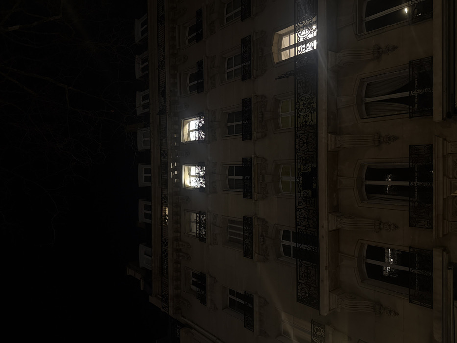 The image displays a multi-story Haussmannian-style apartment building facade at night in Paris, France. The structure, constructed of light-colored stone, features multiple levels of windows, many adorned with ornate black wrought iron balconies. Decorative corbels support several balconies. The upper portion of the building includes a visible mansard roof with dormer windows. The scene is predominantly dark, illuminated primarily by interior lights emanating from four windows on two different floors. Two adjacent windows on an upper floor are brightly lit, revealing internal curtains or blinds. Another window on the same floor, to the far right, is also illuminated. A window on a lower floor, also to the far right, shows similar internal lighting. No people, specific actions, or interactions are visible. Bare tree branches are discernible against the dark night sky in the upper right of the frame.