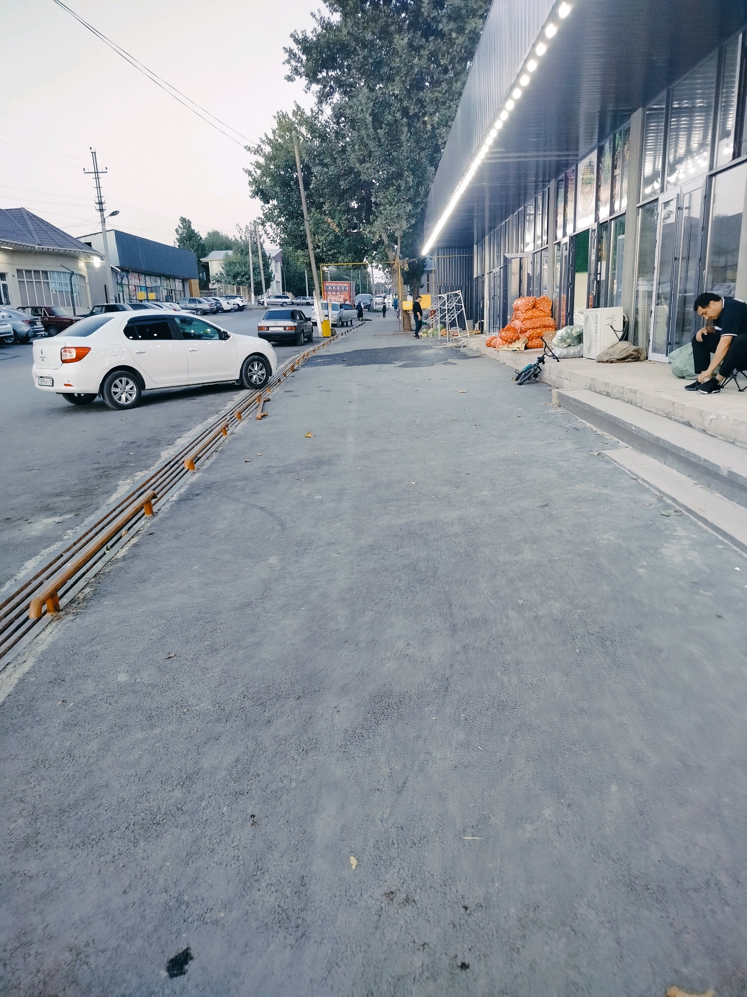 The image depicts a street scene in the city of Kölkent, Kazakhstan, likely in front of a market or commercial building. A white sedan is parked on the asphalt road. In the background, several other vehicles are visible, along with a building featuring a modern facade with large glass windows and an overhang illuminated by a string of lights. Piles of produce, possibly onions, and other goods are stacked on the sidewalk. A bicycle is leaning against a rack. A man in a black shirt and pants is seated on a step, attending to his shoes. The environment appears to be outdoors, with trees lining the street and a power pole visible. The sky is overcast, suggesting either early morning or late evening.