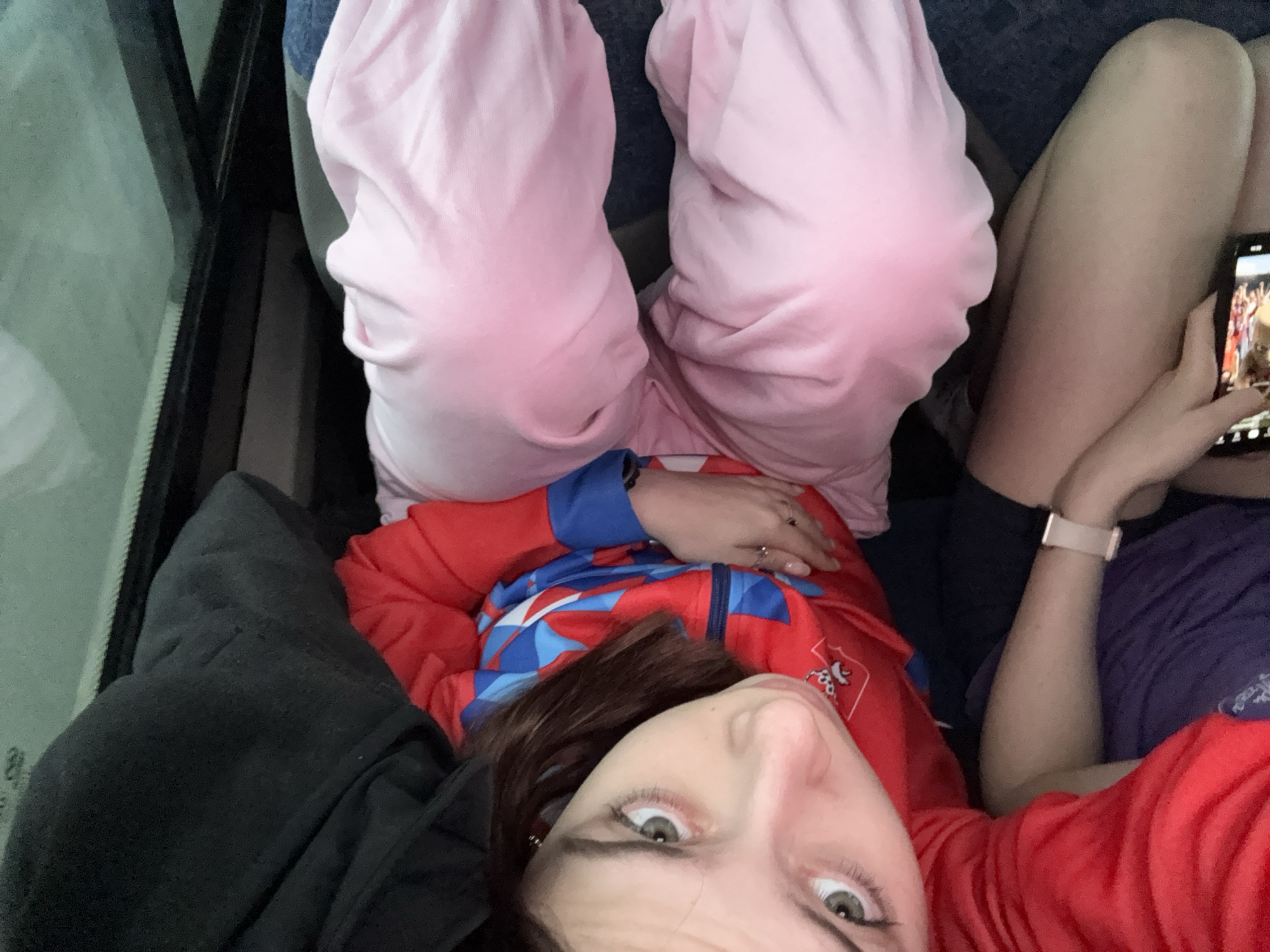 A low-angle, close-up shot shows a person with fair skin and light-colored eyes lying down. The person is wearing a red and blue zip-up jacket. Above them are pink sweatpants. Beside the person, another individual is holding a smartphone displaying a basketball game. The scene appears to be inside a vehicle. There are indications that the picture may have been taken in the city of Perm, Russia because of the person’s sweatshirt.