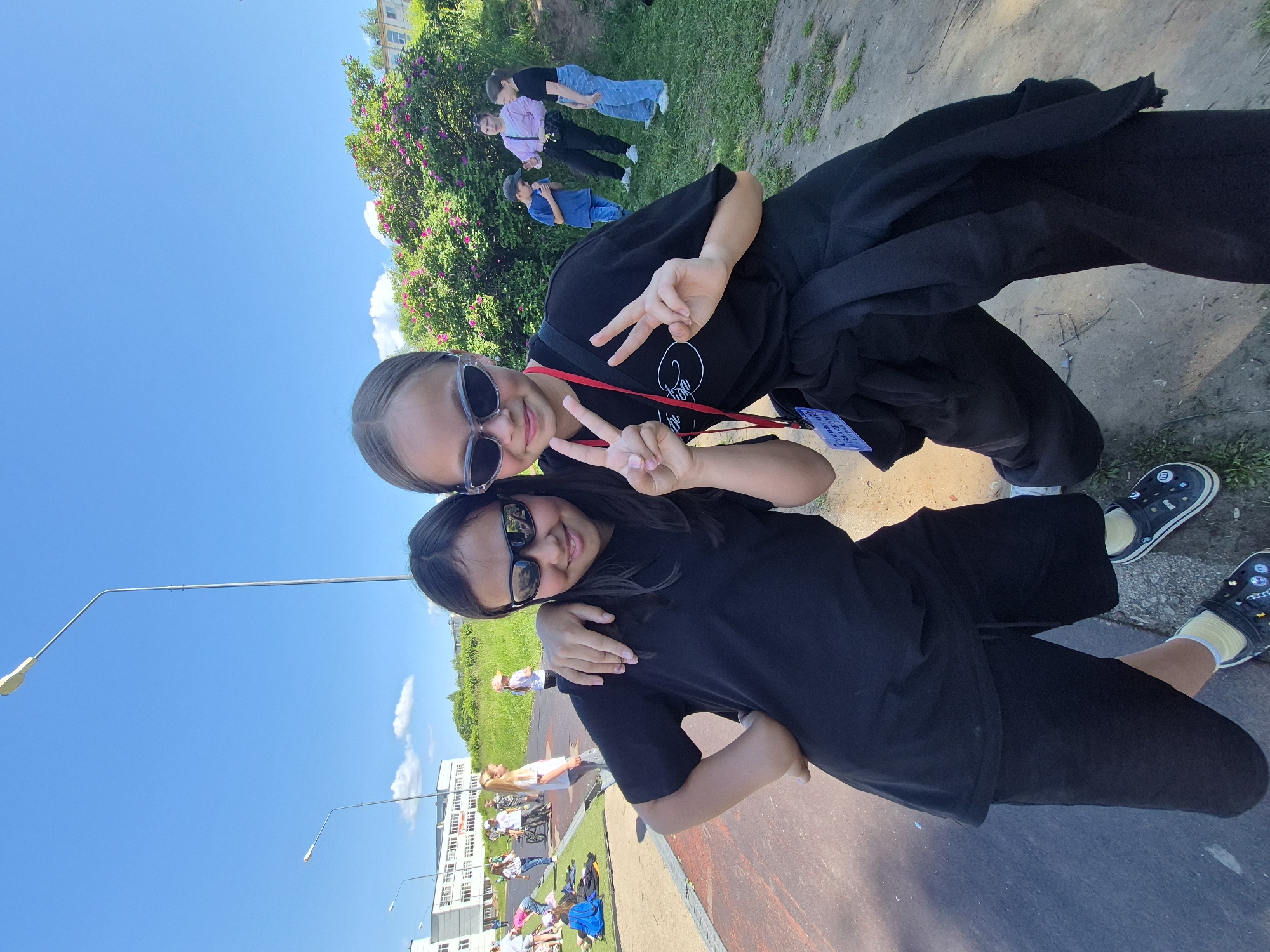 The image shows two young girls posing for a photo outdoors in Chaikovsky, Russia. The girl on the left has dark hair, is wearing sunglasses, a black t-shirt, and black shorts. The girl on the right also wears sunglasses and a black t-shirt and is making a peace sign. Behind them, other children and adults are visible in the background, some gathered near a flowering bush, while others are on a grassy area and near a building that appears to be a school. The setting seems to be a park or recreational area, with a street lamp and a building in the distance. The sky is clear and blue, suggesting a sunny day.
