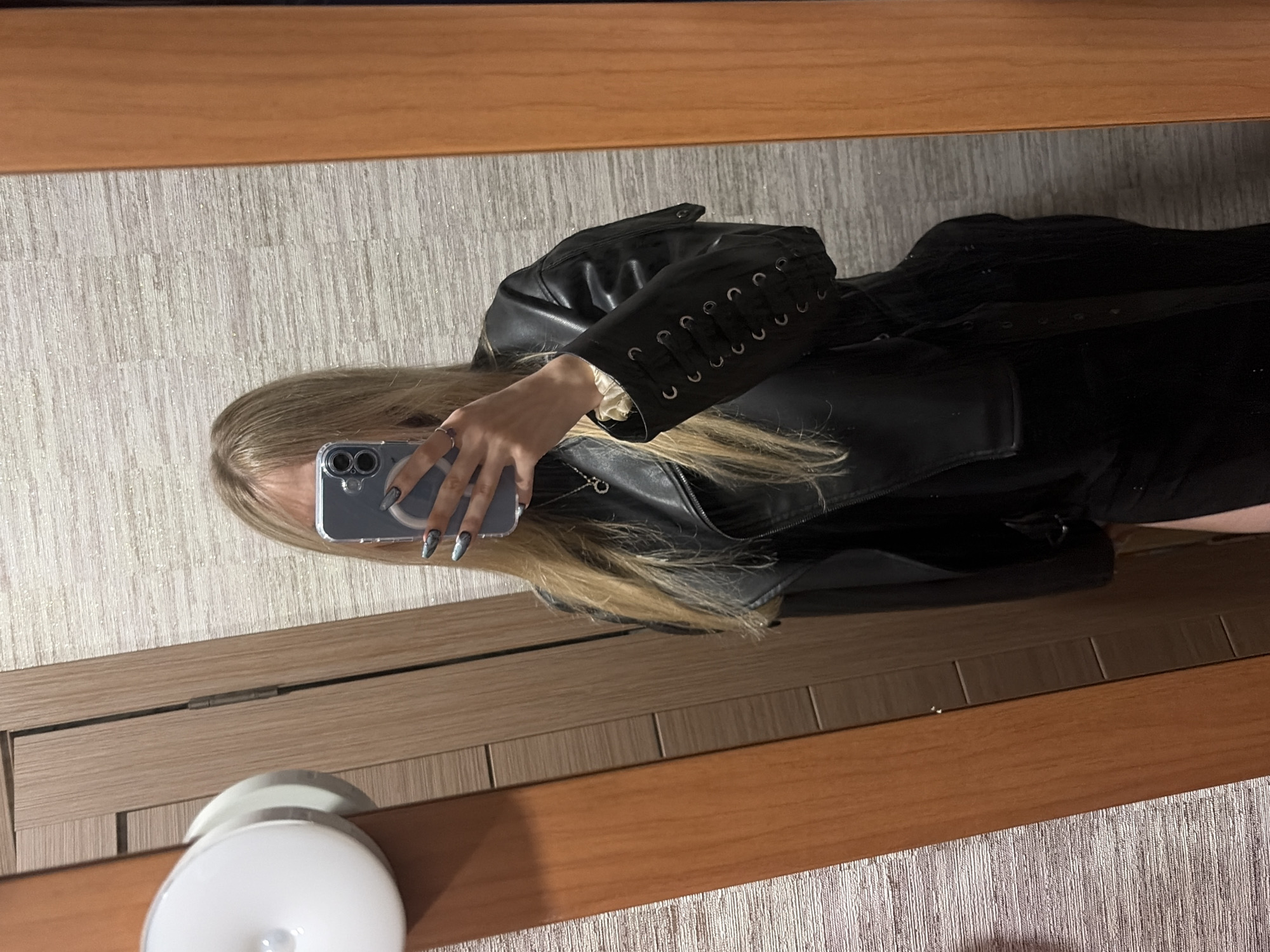 A woman with long blonde hair is holding a blue iPhone to take a selfie in a mirror. She is wearing a black leather jacket with decorative lacing on the sleeve, and a black dress or top. Her nails are long and painted with a dark, intricate design. The mirror is framed with a light brown wood, and there is a wall with a subtle vertical pattern behind her. A partially visible white object, possibly a lamp or a decorative item, is in the foreground. The location is Unknown, Unknown.