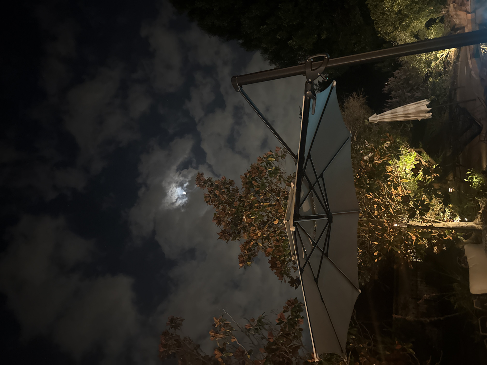 At night in Southlake, United States of America, an upward shot focuses on a partially opened gray patio umbrella. The umbrella's support pole extends out from the top right, with some hardware attached. The night sky is partially obscured by clouds, with a glimpse of the moon visible. Foliage fills the right side of the frame, some lit by an unseen light source. A closed, striped umbrella is also visible in the foliage.