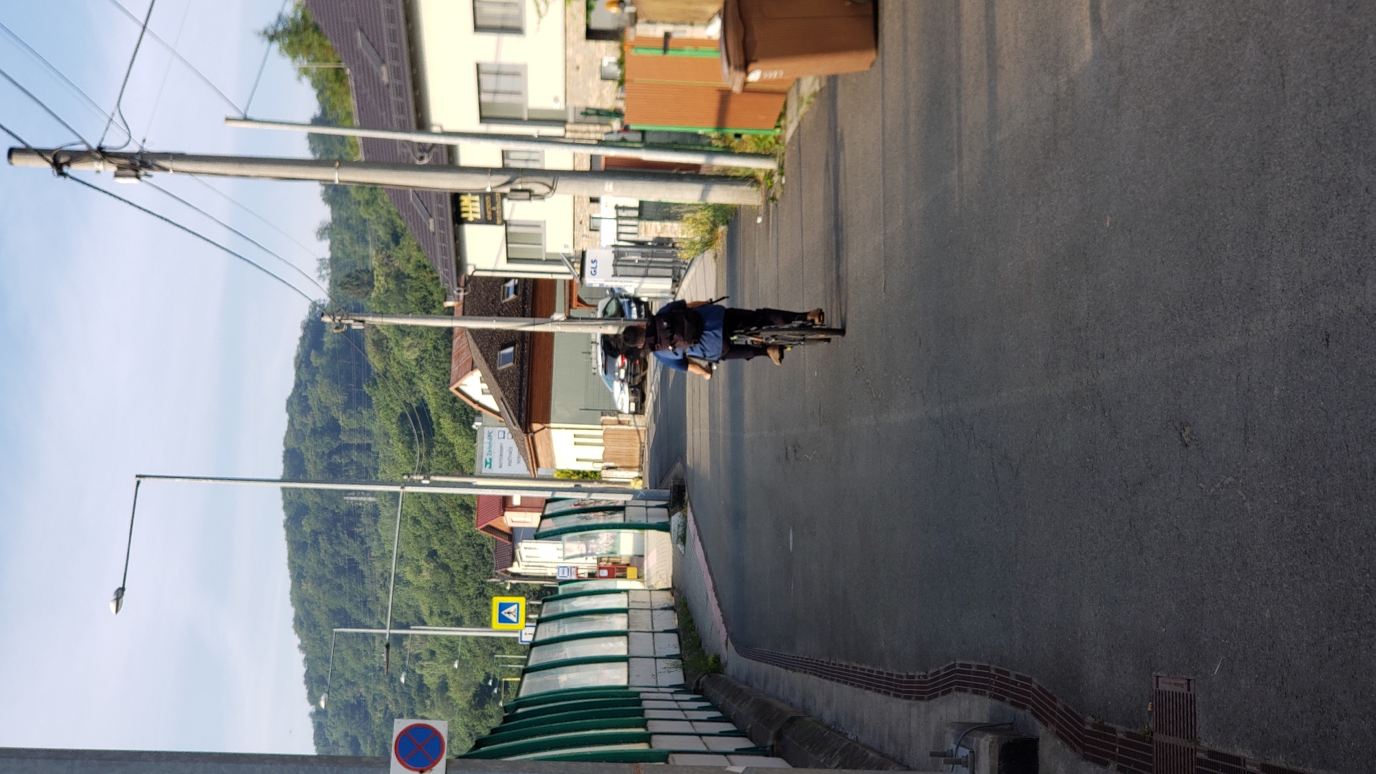 A person on a bicycle rides down a paved street in Prešov, Slovakia. The cyclist, seen from behind, wears a dark t-shirt, dark pants, and a black backpack. They are riding a dark-colored bicycle. The street is flanked by buildings and a green and white sound barrier on the left side. In the background, a lush green hill rises under a bright sky with scattered clouds. There are streetlights and utility poles along the street. A brown trash bin is visible on the right side of the image. The scene appears to be in a residential or semi-commercial area.